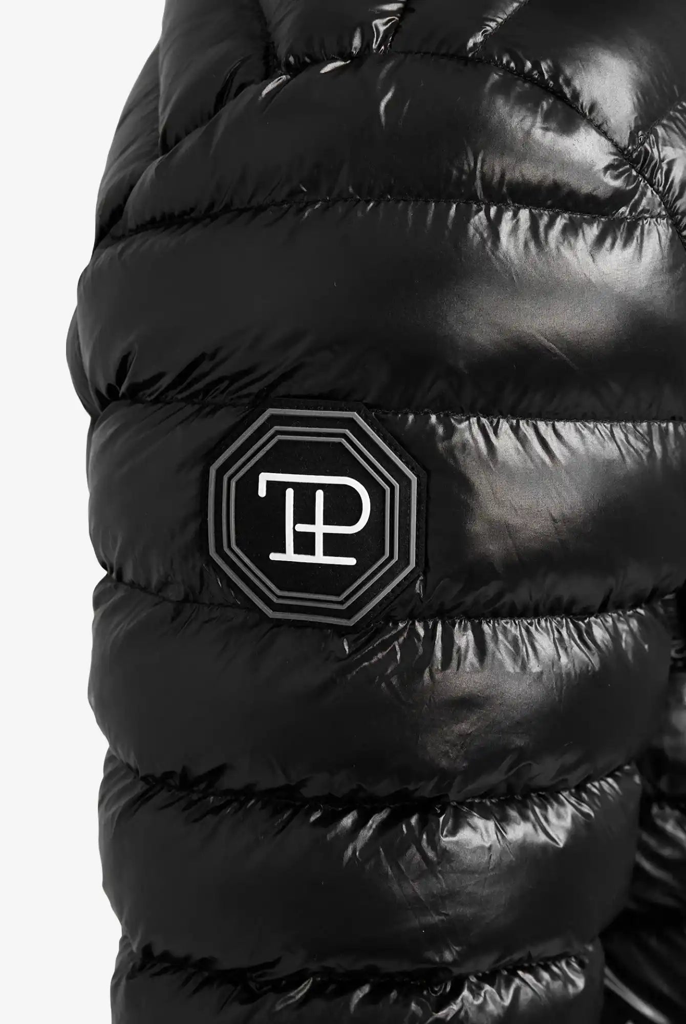 Eine Nahaufnahme einer glänzend schwarzen Adan Steppjacke mit Kapuze von trueprodigy zeigt einen wasserabweisenden Ärmel mit einem achteckigen schwarzen Aufnäher, auf dem weiße geometrische Buchstaben gestickt sind.
