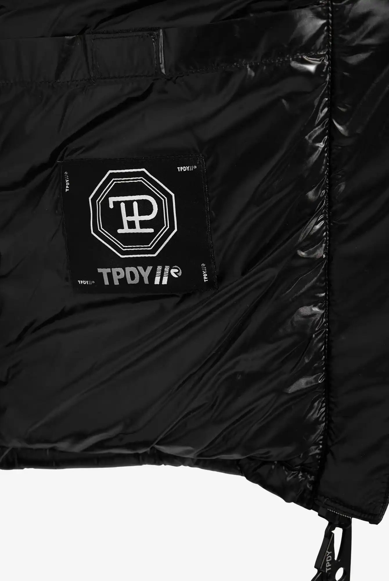 Nahaufnahme der Innenseite der glänzenden, wasserabweisenden trueprodigy Adan Steppjacke mit Kapuze, die einen aufgenähten schwarzen Aufnäher mit weißen geometrischen und TPDY-Textmotiven zeigt. Die gesteppte Textur und die Kapuze sorgen für Stil.