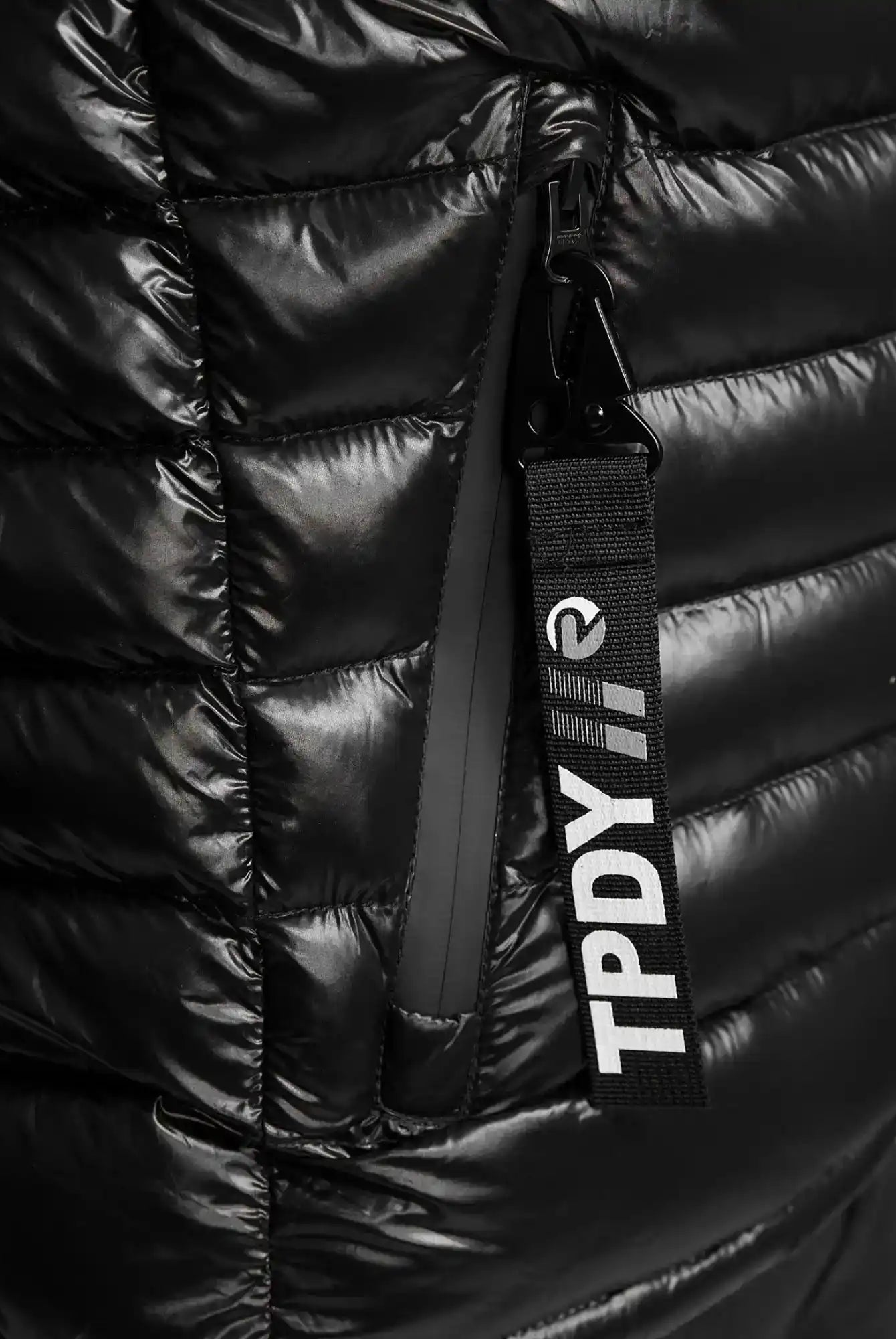 Nahaufnahme einer schwarzen, glänzenden, gesteppten Adan Steppjacke mit Kapuze von trueprodigy. Mit einer vertikalen Reißverschlusstasche und einem schwarzen Anhänger mit weißer Aufschrift "TPDY//".