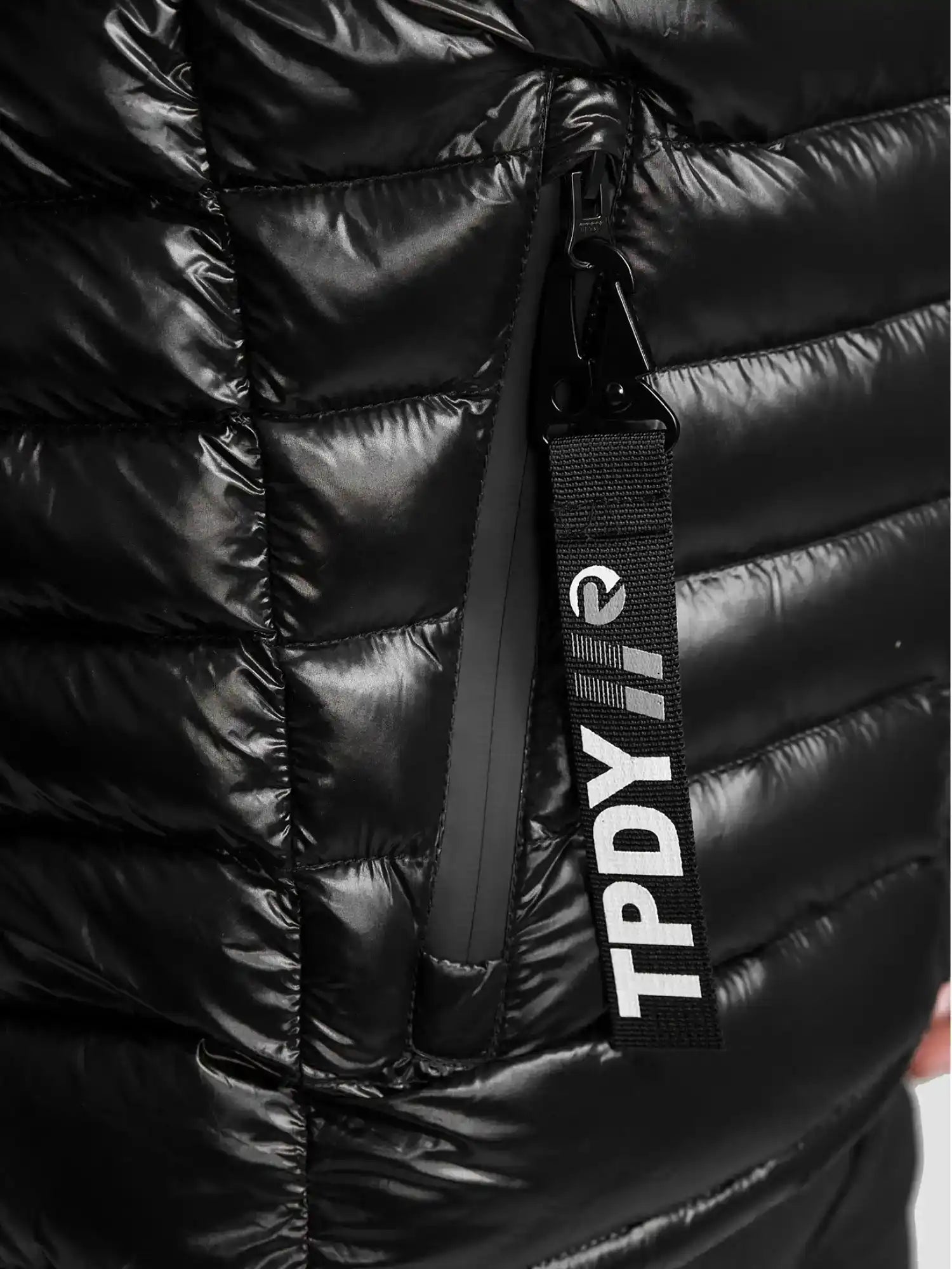 Nahaufnahme einer schwarzen, glänzenden, gesteppten Adan Steppjacke mit Kapuze von trueprodigy. Mit einer vertikalen Reißverschlusstasche und einem schwarzen Anhänger mit weißer Aufschrift "TPDY//".
