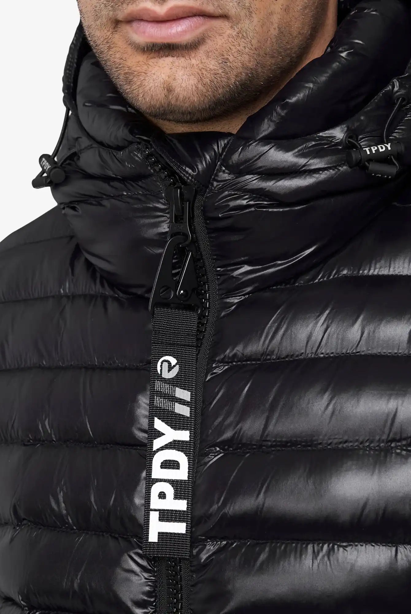Nahaufnahme einer Person, die die glänzende schwarze Adan Steppjacke mit Kapuze von trueprodigy trägt. Die wasserabweisende Jacke ist mit einem großen TPDY-Reißverschluss in Weiß versehen, und ein Teil der Unterseite ist sichtbar.