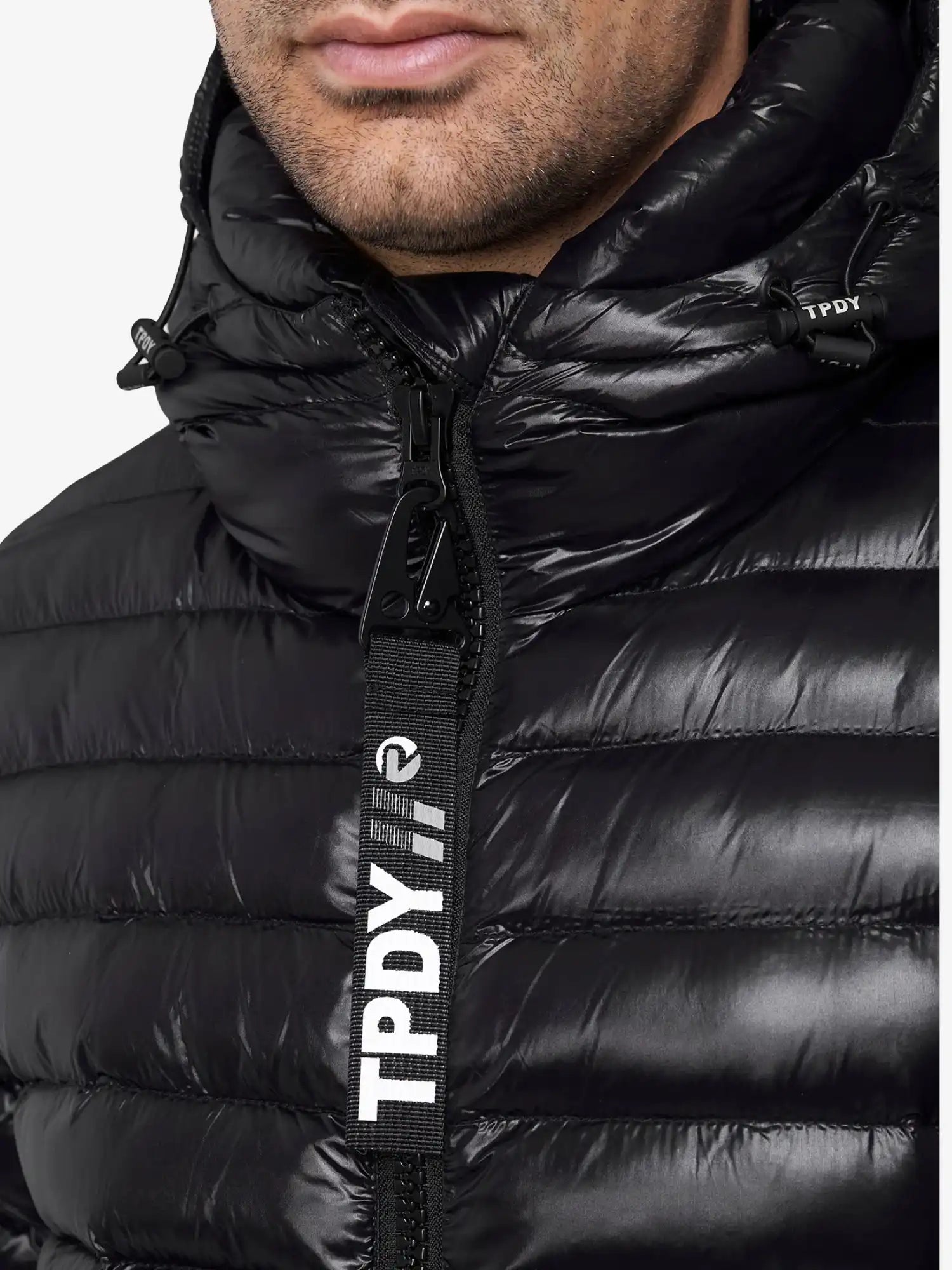 Nahaufnahme einer Person, die die glänzende schwarze Adan Steppjacke mit Kapuze von trueprodigy trägt. Die wasserabweisende Jacke ist mit einem großen TPDY-Reißverschluss in Weiß versehen, und ein Teil der Unterseite ist sichtbar.
