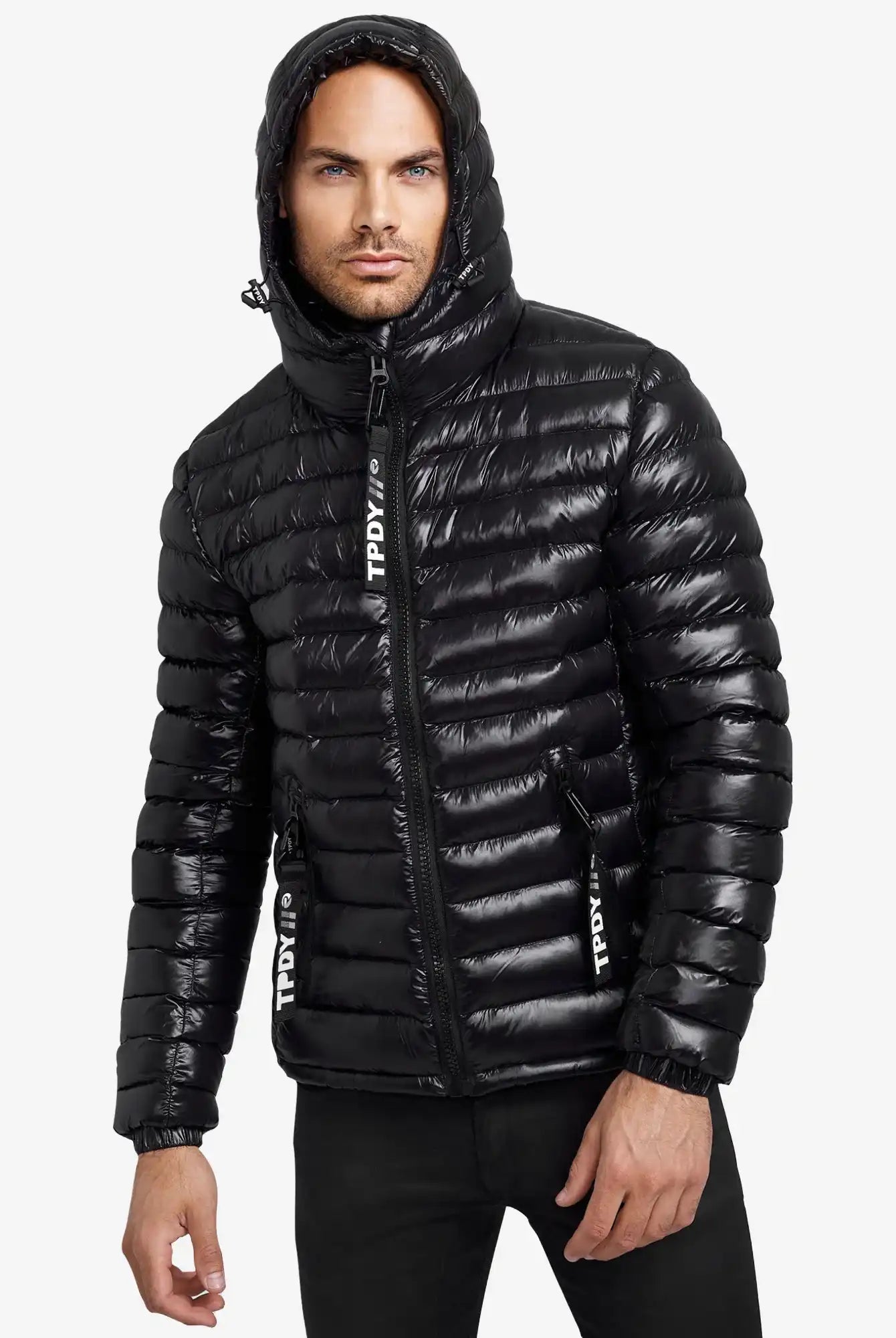 Ein Mann trägt die Adan Steppjacke mit Kapuze von trueprodigy-eine schwarze, wasserabweisende Herrenjacke mit weißen Schriftzügen an den Reißverschlüssen. Er steht vor weißem Hintergrund und blickt seitlich neutral.