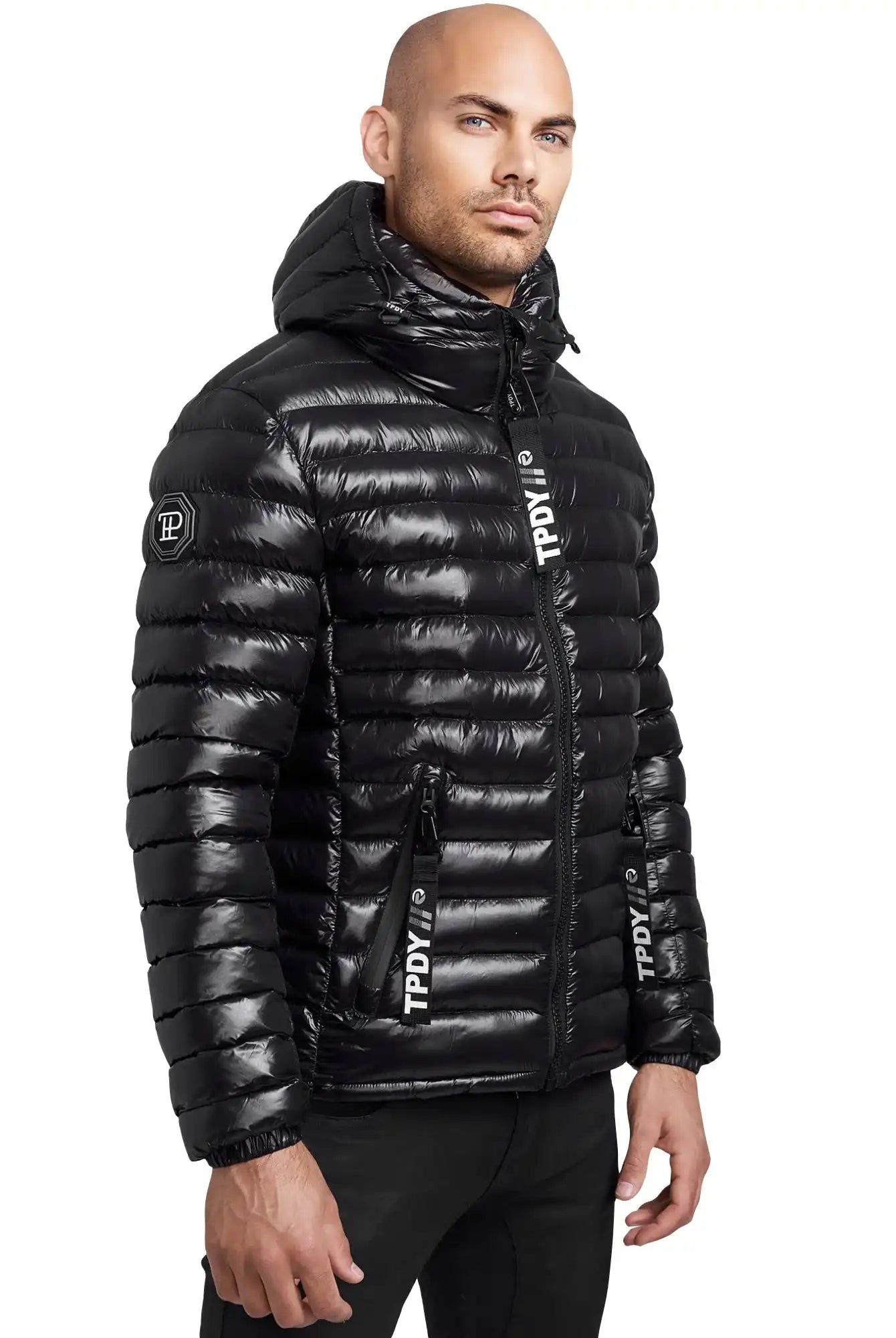 Ein Mann trägt eine glänzende schwarze Trueprodigy Adan Steppjacke mit Kapuze, die leicht umgeschlagen ist. Die wasserabweisende Jacke hat horizontale Steppungen, Reißverschlusstaschen und Logotext auf dem Ärmel, der Brust und der Reißverschlussklappe.