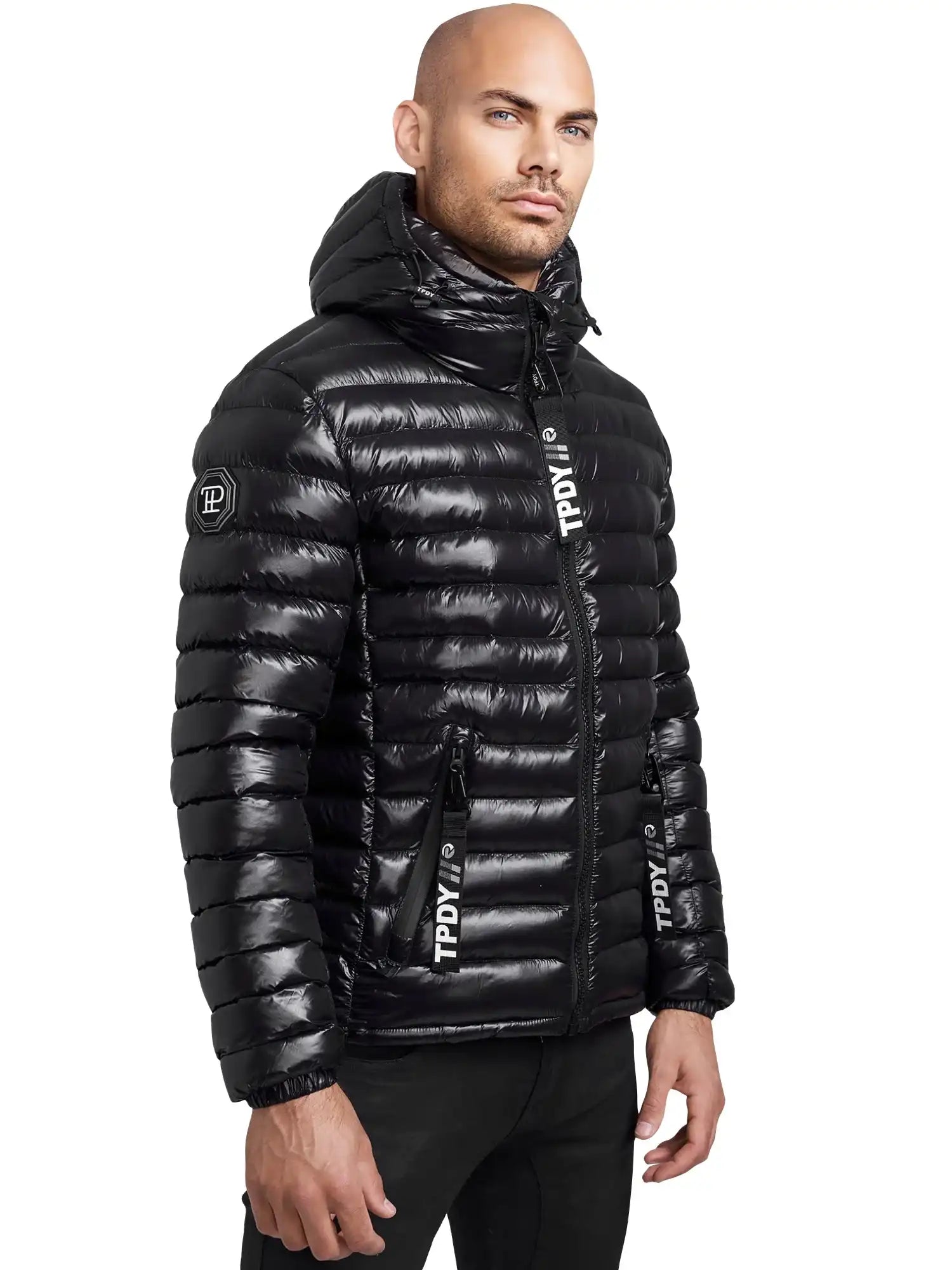 Ein Mann trägt eine glänzende schwarze Trueprodigy Adan Steppjacke mit Kapuze, die leicht umgeschlagen ist. Die wasserabweisende Jacke hat horizontale Steppungen, Reißverschlusstaschen und Logotext auf dem Ärmel, der Brust und der Reißverschlussklappe.
