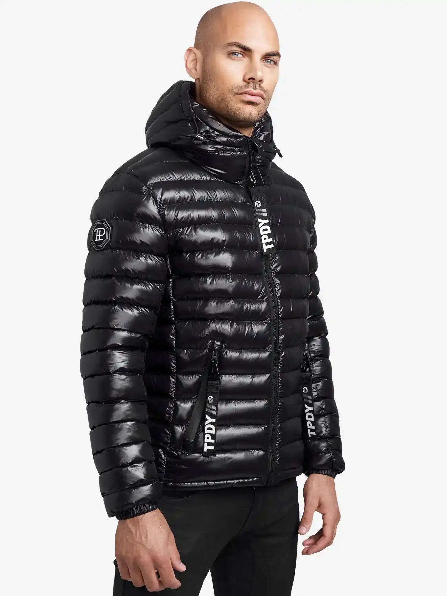 Ein Mann mit rasiertem Kopf und kurzem Bart trägt die glänzende, wasserabweisende trueprodigy Adan Steppjacke mit Kapuze mit TPPLY-Branding auf der Vorderseite und dem Ärmel. Er steht vor einem schlichten weißen Hintergrund.