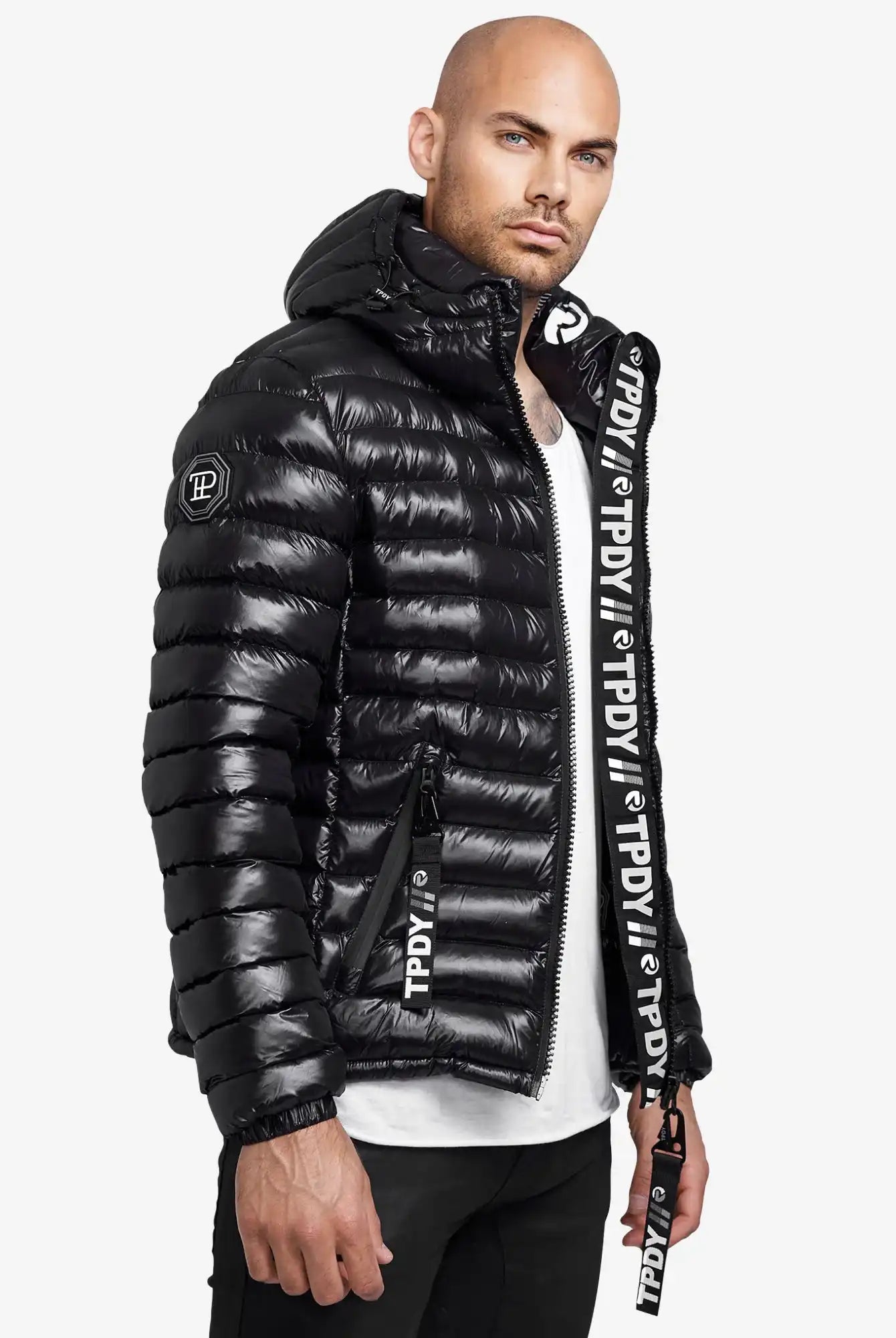 Ein Mann in einer glänzenden schwarzen Adan Steppjacke mit Kapuze von trueprodigy, weißem T-Shirt und schwarzer Hose steht leicht zur Seite gedreht. Die Jacke hat einen auffälligen weißen Schriftzug an den Reißverschlüssen und der Zierleiste.