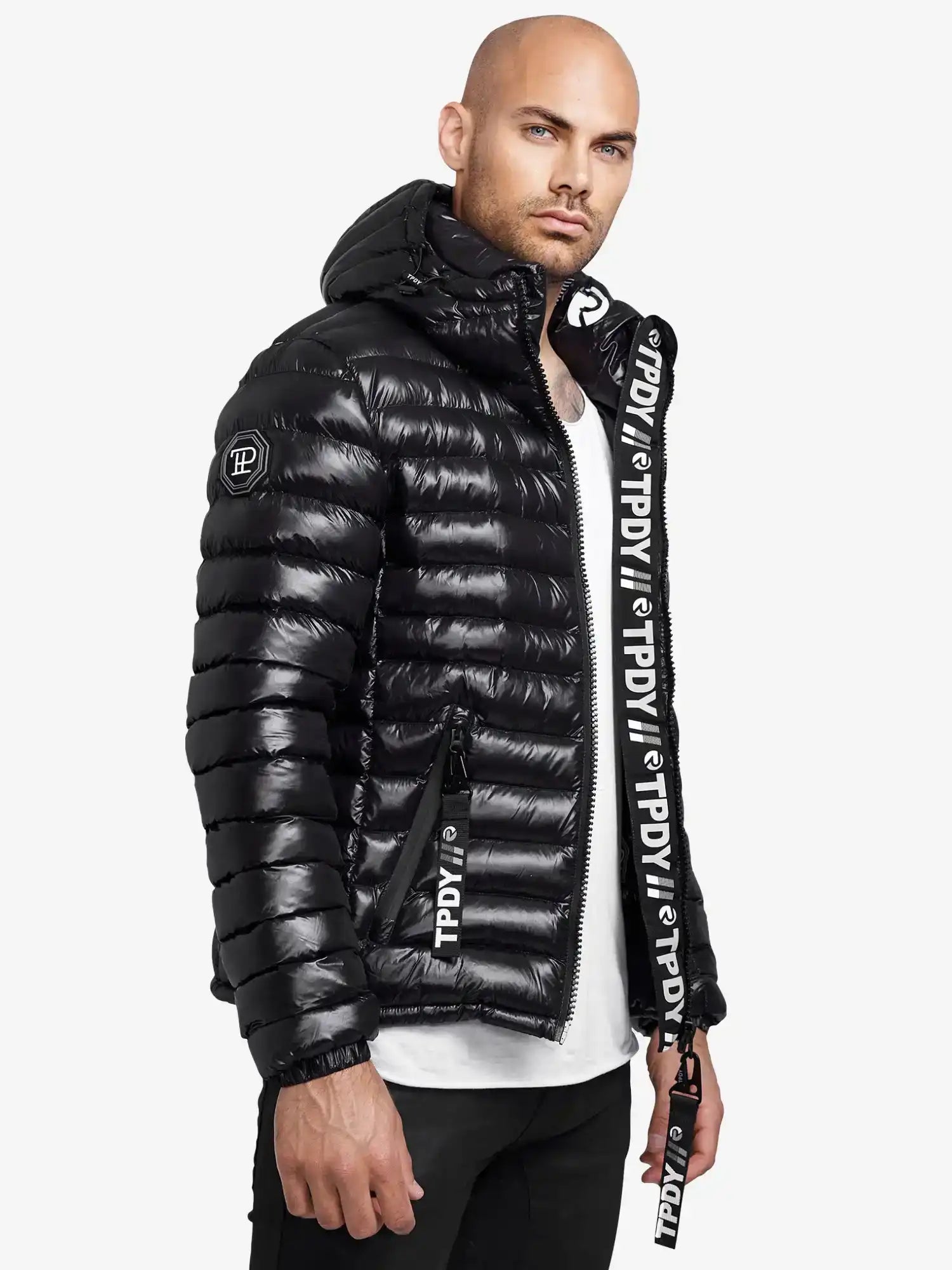 Ein Mann in einer glänzenden schwarzen Adan Steppjacke mit Kapuze von trueprodigy, weißem T-Shirt und schwarzer Hose steht leicht zur Seite gedreht. Die Jacke hat einen auffälligen weißen Schriftzug an den Reißverschlüssen und der Zierleiste.