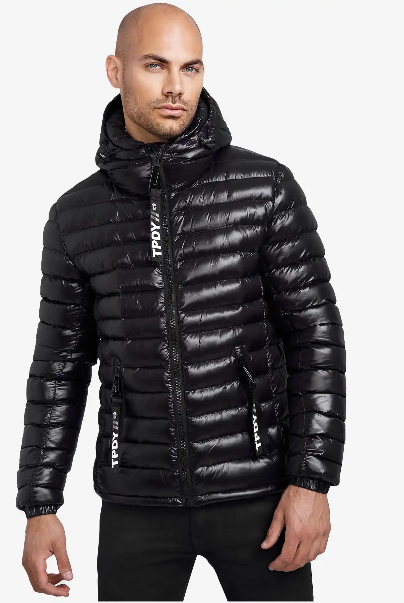 Ein Mann blickt nach vorne in einer glänzend schwarzen trueprodigy Adan Steppjacke mit Kapuze, die mit einer Kapuze, einem Reißverschluss auf der Vorderseite, einer wasserabweisenden Ausrüstung und weißem TPDIVI-Text auf den Reißverschlussleisten vor einem schlichten weißen Hintergrund ausgestattet ist.
