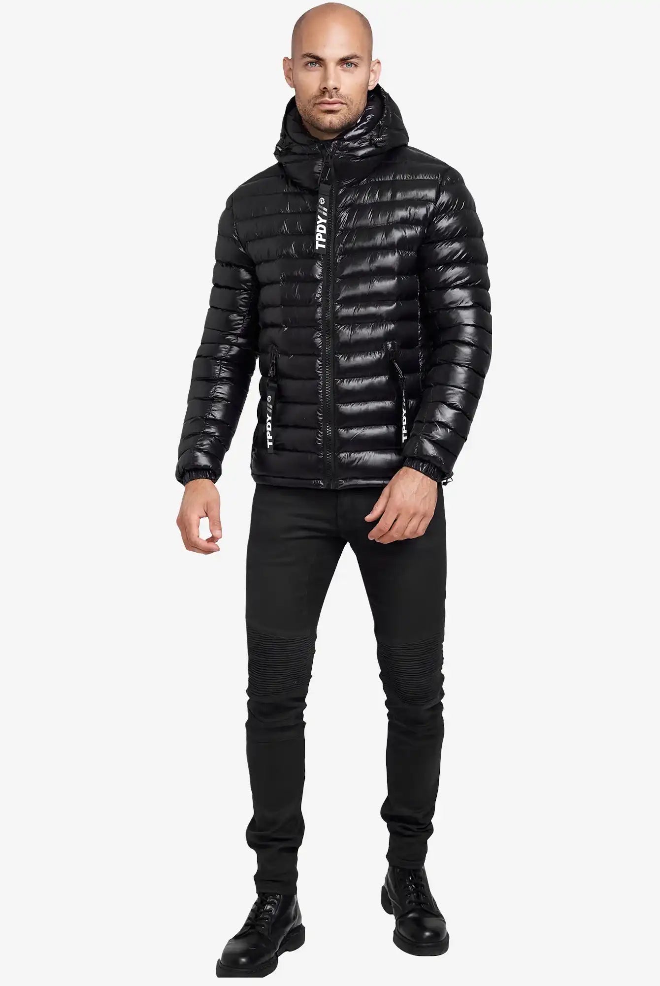 Ein Mann steht mit Blick nach vorne und trägt die glänzende, wasserabweisende schwarze "Adan Steppjacke mit Kapuze" von trueprodigy, gepaart mit einer schmal geschnittenen schwarzen Jeans und schwarzen Schnürstiefeln vor einem schlichten weißen Hintergrund.