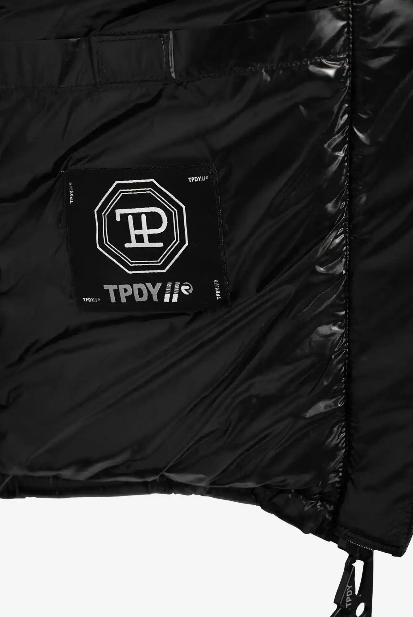 Nahaufnahme der Innenseite der trueprodigy Adan F Steppjacke mit Kapuze, die ein aufgenähtes schwarzes Stoffetikett mit weißer Grafik und "TPDY//"-Text sowie geometrische Muster auf einem glänzenden schwarzen Futter zeigt.