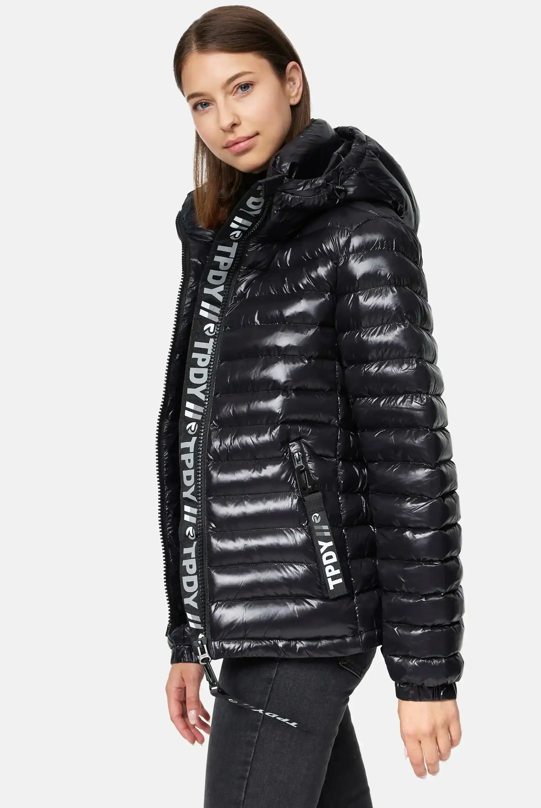 Eine Frau trägt die Adan F Steppjacke mit Kapuze von trueprodigy in glänzendem Schwarz, mit auffälligem Text am Reißverschluss sowie innen. Die wasserabweisende Jacke eignet sich perfekt für jedes Wetter und wird vor weißem Hintergrund gezeigt.