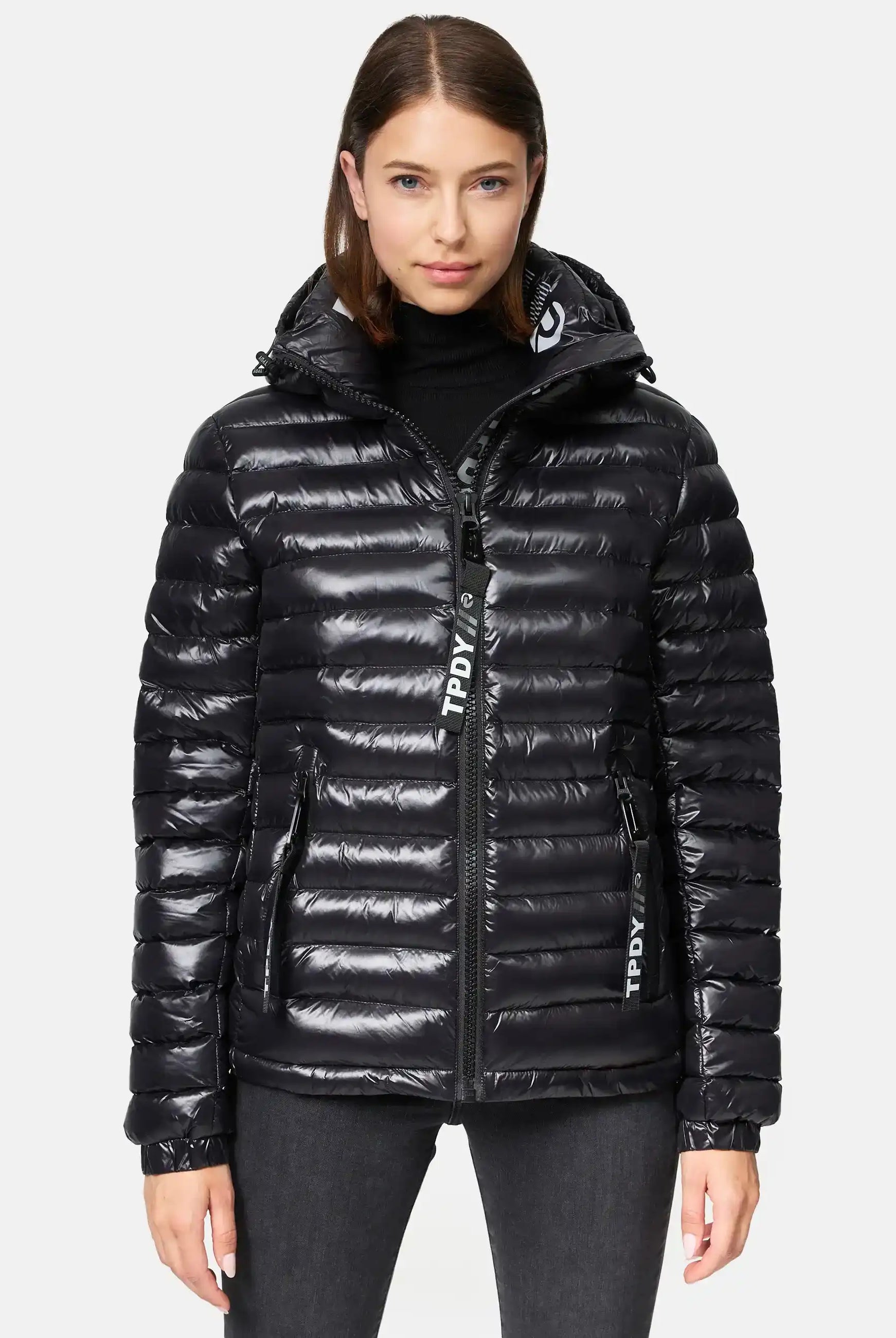 Eine Frau in einer glänzenden schwarzen Adan F Steppjacke mit Kapuze von trueprodigy mit Frontreißverschluss und zwei Seitentaschen steht vor einem weißen Hintergrund und blickt direkt in die Kamera. Die Jacke ist wasserabweisend.