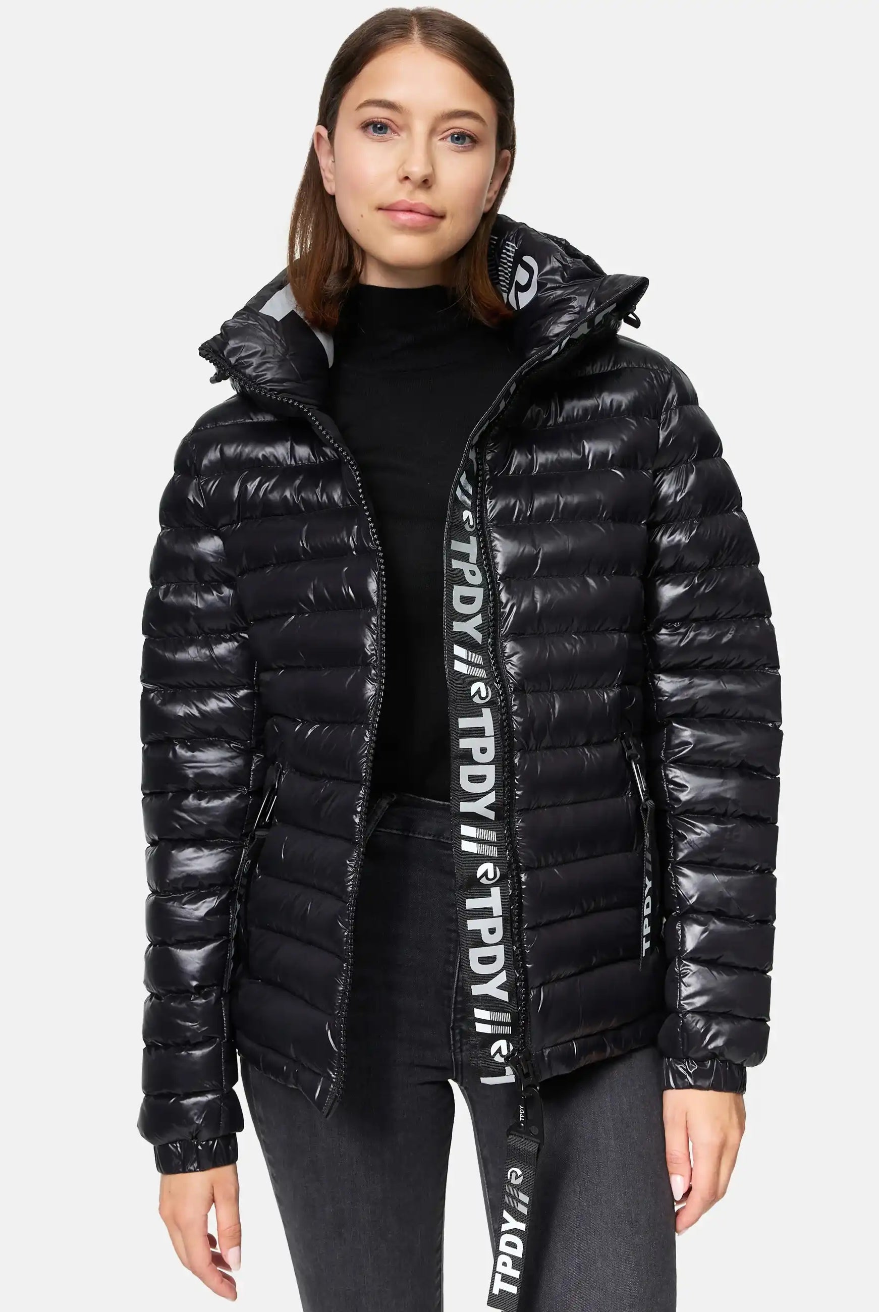 Eine Frau blickt nach vorne in einer glänzenden schwarzen trueprodigy Adan F Steppjacke mit Kapuze über einem schwarzen Rollkragenpullover und dunklen Jeans. Die Jacke hat ein Logoband, sichtbaren Reißverschluss und ist wasserabweisend. Der Hintergrund ist einfarbig und hell.