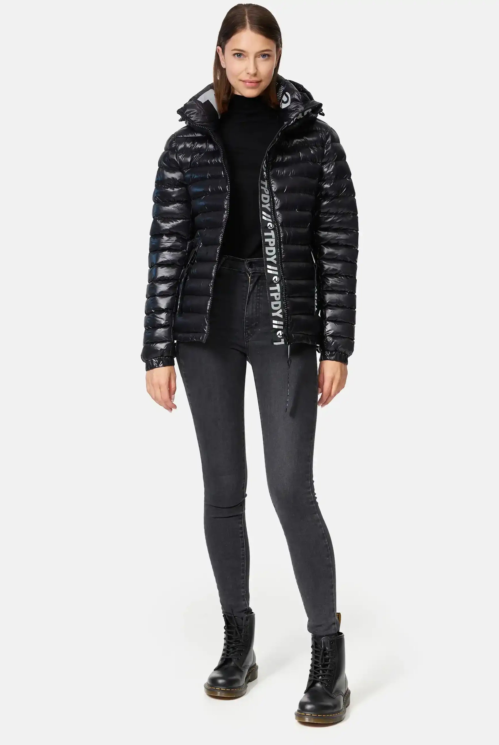 Eine Frau blickt nach vorne und trägt eine glänzende, schwarze Trueprodigy Adan F Steppjacke mit Kapuze, einen schwarzen Rollkragenpullover, dunkle, hochgeschnittene Skinny Jeans und schwarze Schnürstiefel; ihr glattes Haar ist in der Mitte gescheitelt.