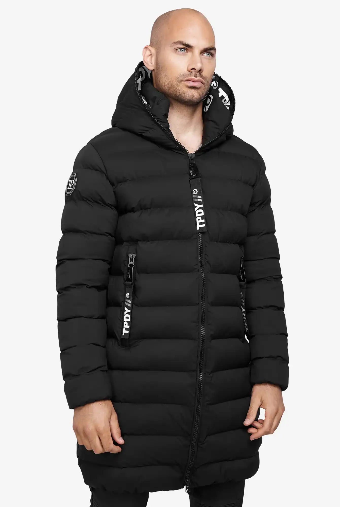 Ein Mann trägt die Chester Lange Pufferjacke mit Logo von trueprodigy im Herrenparka-Stil. Die schwarze, wasserabweisende Jacke hat sichtbare Reißverschlüsse sowie weiße Logodetails an Kapuze und Zipper.