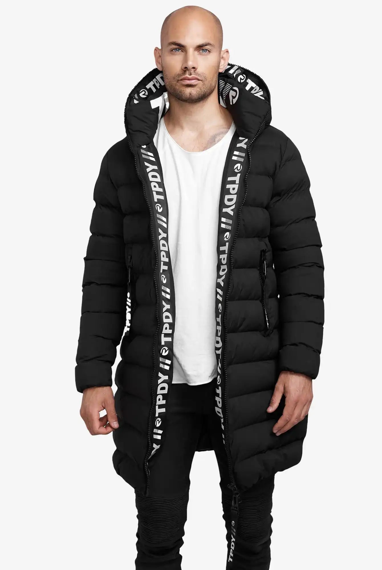 Ein Mann steht vor der Kamera und trägt die trueprodigy Chester Lange Pufferjacke mit Logo mit Kapuze und weißem Logo-Schriftzug am Saum, gepaart mit einem weißen T-Shirt und einer schwarzen Hose vor einem schlichten weißen Hintergrund.