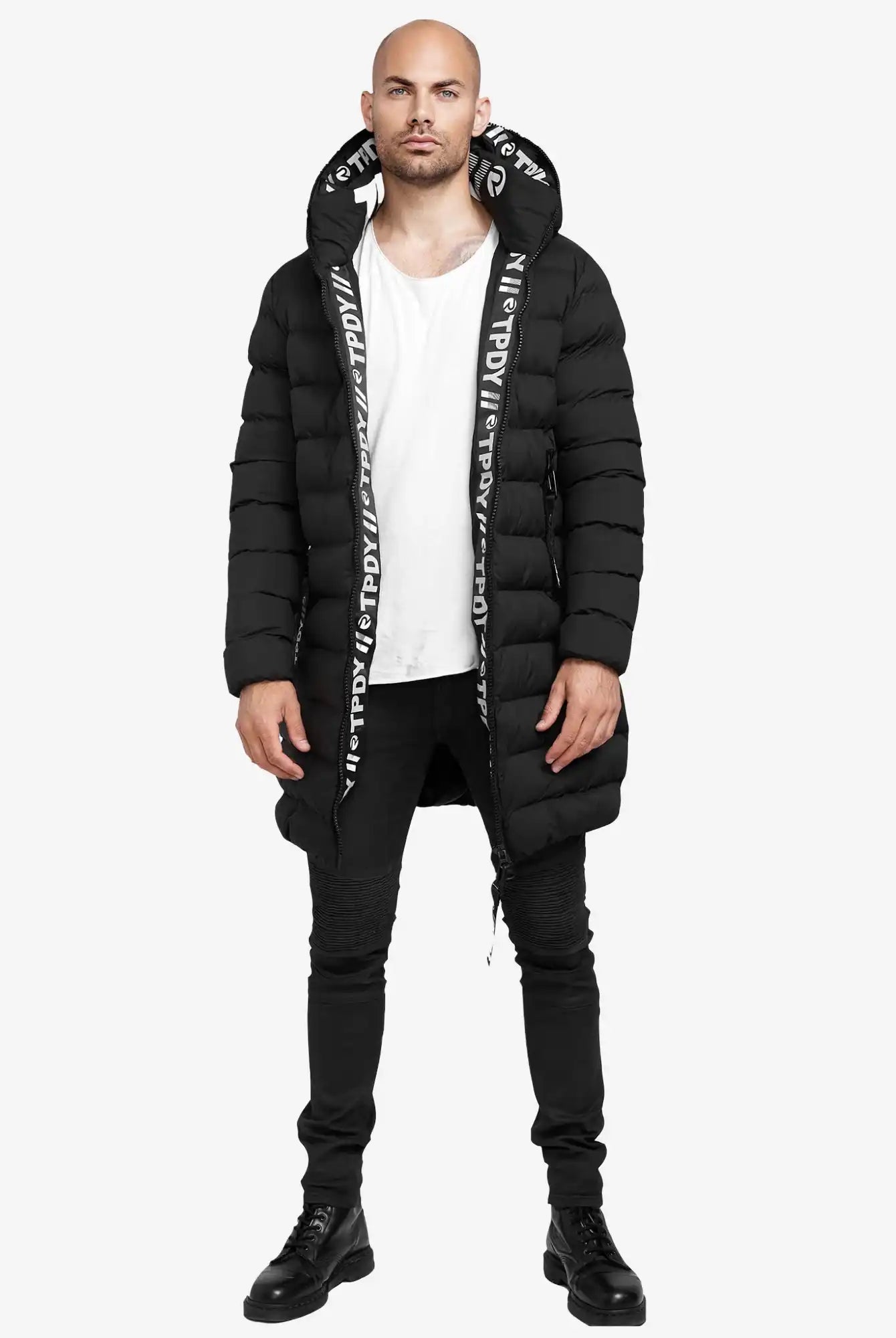 Ein Mann trägt die Chester Lange Pufferjacke mit Logo von trueprodigy in Schwarz mit Kapuze, kombiniert mit weißem T-Shirt, schwarzen Skinny-Jeans und Boots. Die Jacke ist wasserabweisend und zeigt ein Logo auf dem Kapuzenfutter.