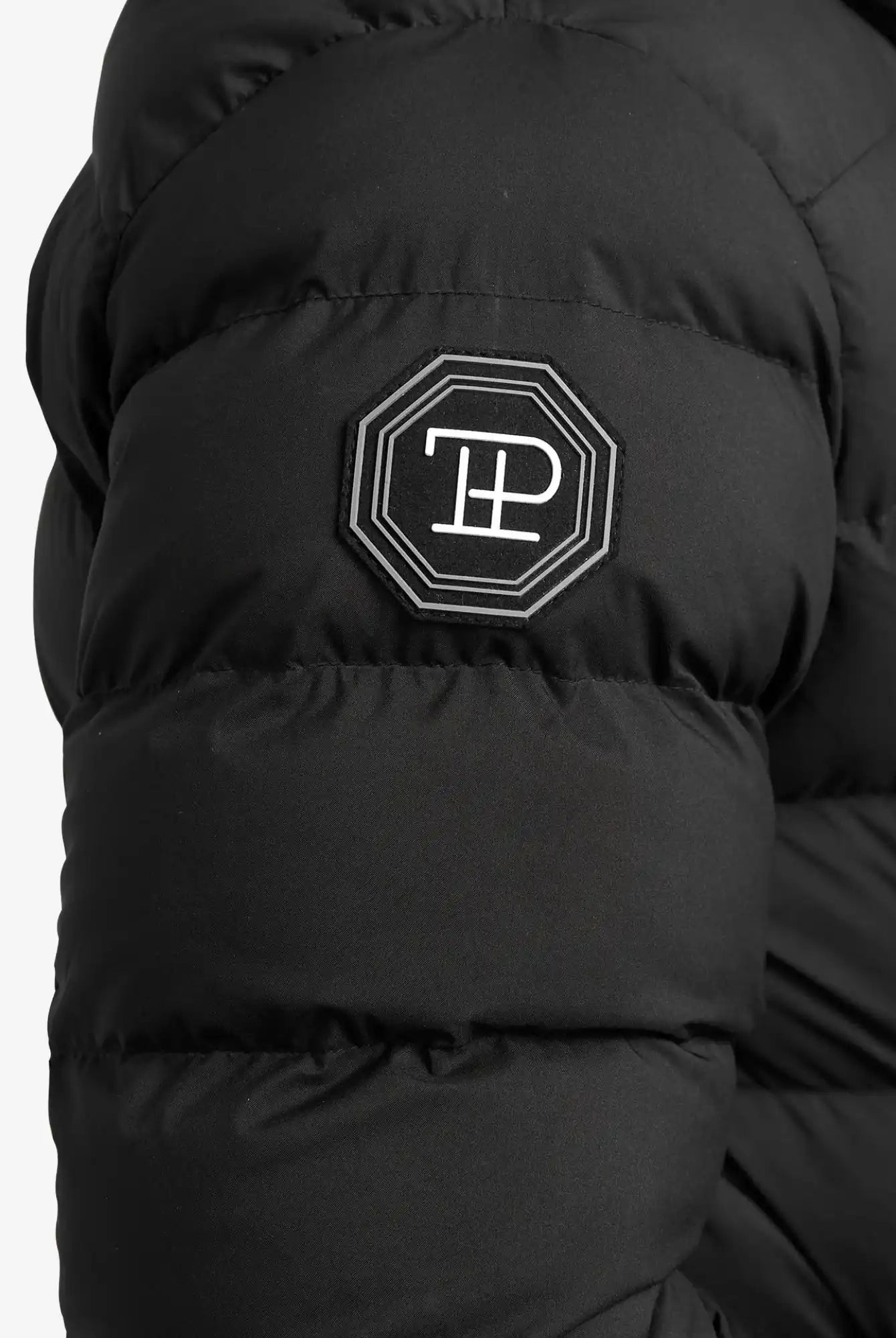 Nahaufnahme der Chester F Lange Pufferjacke mit Logo von trueprodigy: schwarze, gesteppte Jacke mit wasserabweisender Funktion und schwarz-weißem, achteckigem TP-Patch am Ärmel - perfekt als Damenparka für moderne Styles.