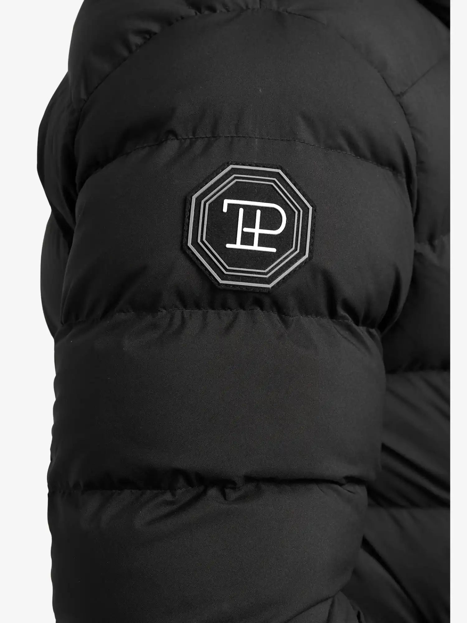 Nahaufnahme der Chester F Lange Pufferjacke mit Logo von trueprodigy: schwarze, gesteppte Jacke mit wasserabweisender Funktion und schwarz-weißem, achteckigem TP-Patch am Ärmel - perfekt als Damenparka für moderne Styles.