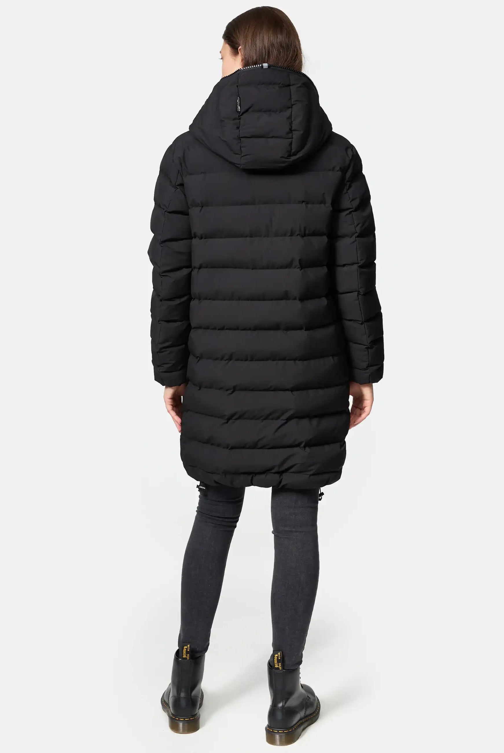 Eine Person steht mit dem Gesicht nach unten und trägt die trueprodigy Chester F Lange Pufferjacke mit Logo, einen langen schwarzen Damenparka mit Kapuze, gepaart mit dunklen Skinny Jeans und schwarzen Stiefeletten mit Reißverschlüssen und Schnallen. Der Hintergrund ist einfarbig weiß.