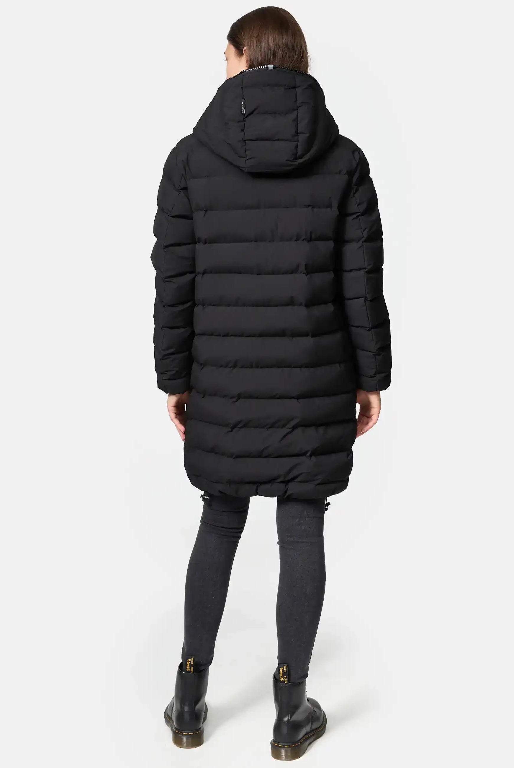 Eine Person steht mit dem Gesicht nach unten und trägt die trueprodigy Chester F Lange Pufferjacke mit Logo, einen langen schwarzen Damenparka mit Kapuze, gepaart mit dunklen Skinny Jeans und schwarzen Stiefeletten mit Reißverschlüssen und Schnallen. Der Hintergrund ist einfarbig weiß.