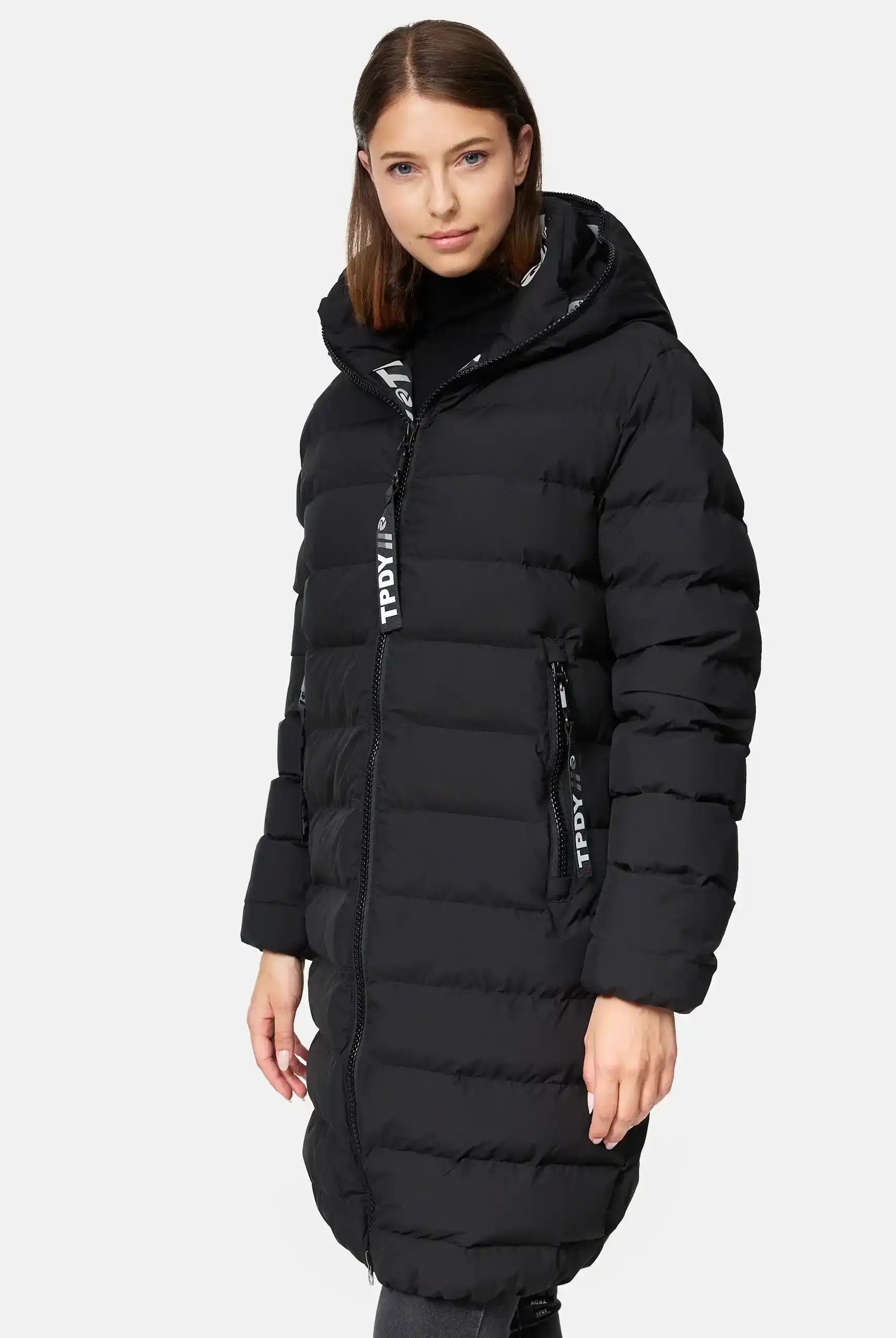 Eine Frau trägt die trueprodigy Chester F Lange Pufferjacke mit Logo, einen schwarzen langen Damenparka mit Kapuze, Frontreißverschluss und seitlichen Reißverschlusstaschen mit gebrandeten Zuglaschen, vor einem schlichten hellen Hintergrund.