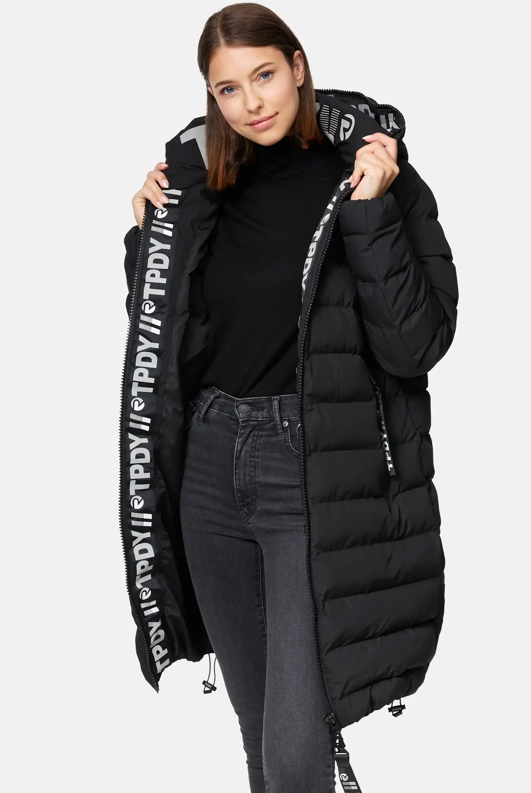 Eine Frau modelliert die trueprodigy Chester F Lange Pufferjacke mit Logo-ein langer, schwarzer, wasserabweisender Damenparka mit auffälligem weißen Logo am Reißverschluss. Sie trägt dazu einen schwarzen Rollkragenpullover und dunkle Jeans.