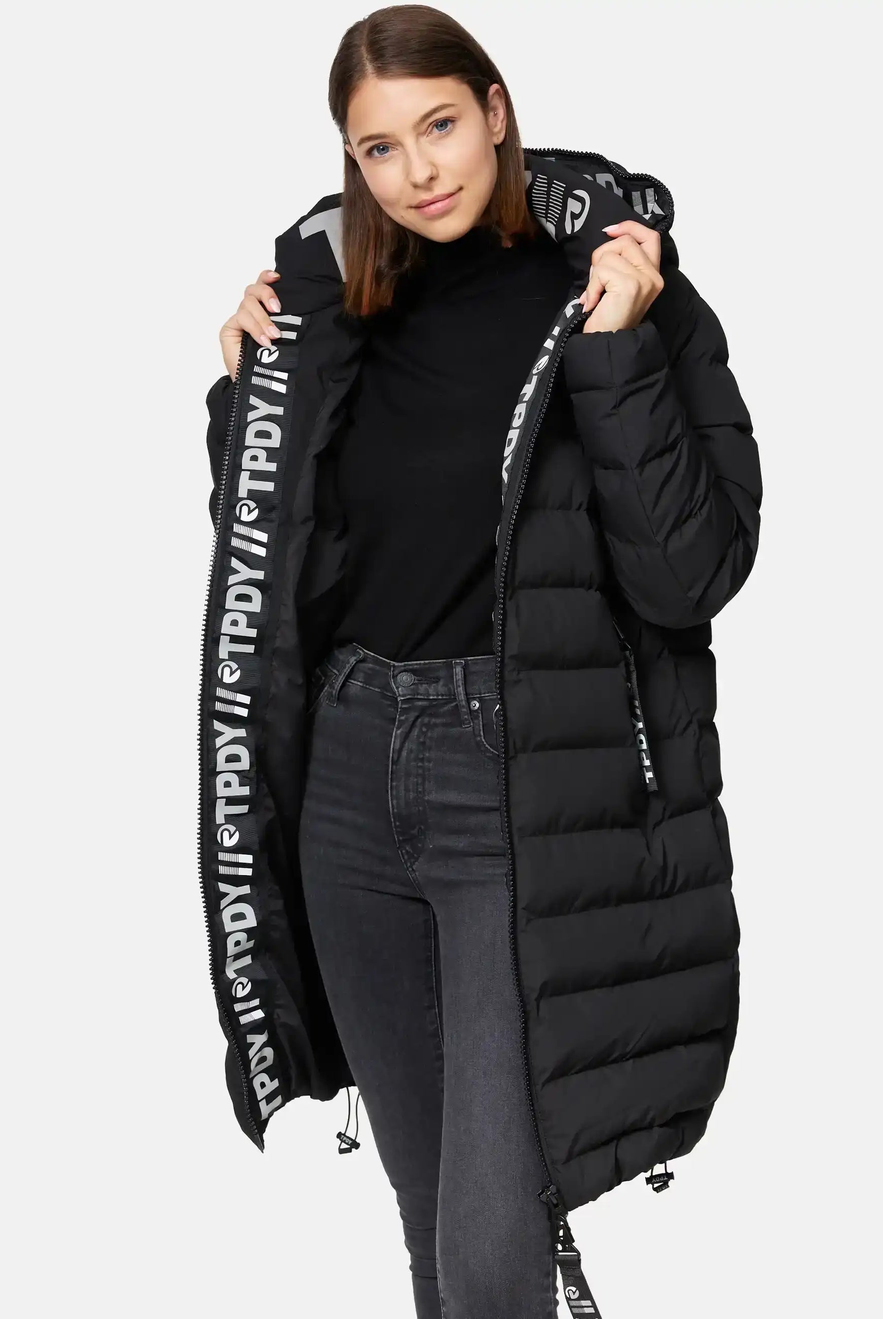 Eine Frau modelliert die trueprodigy Chester F Lange Pufferjacke mit Logo-ein langer, schwarzer, wasserabweisender Damenparka mit auffälligem weißen Logo am Reißverschluss. Sie trägt dazu einen schwarzen Rollkragenpullover und dunkle Jeans.