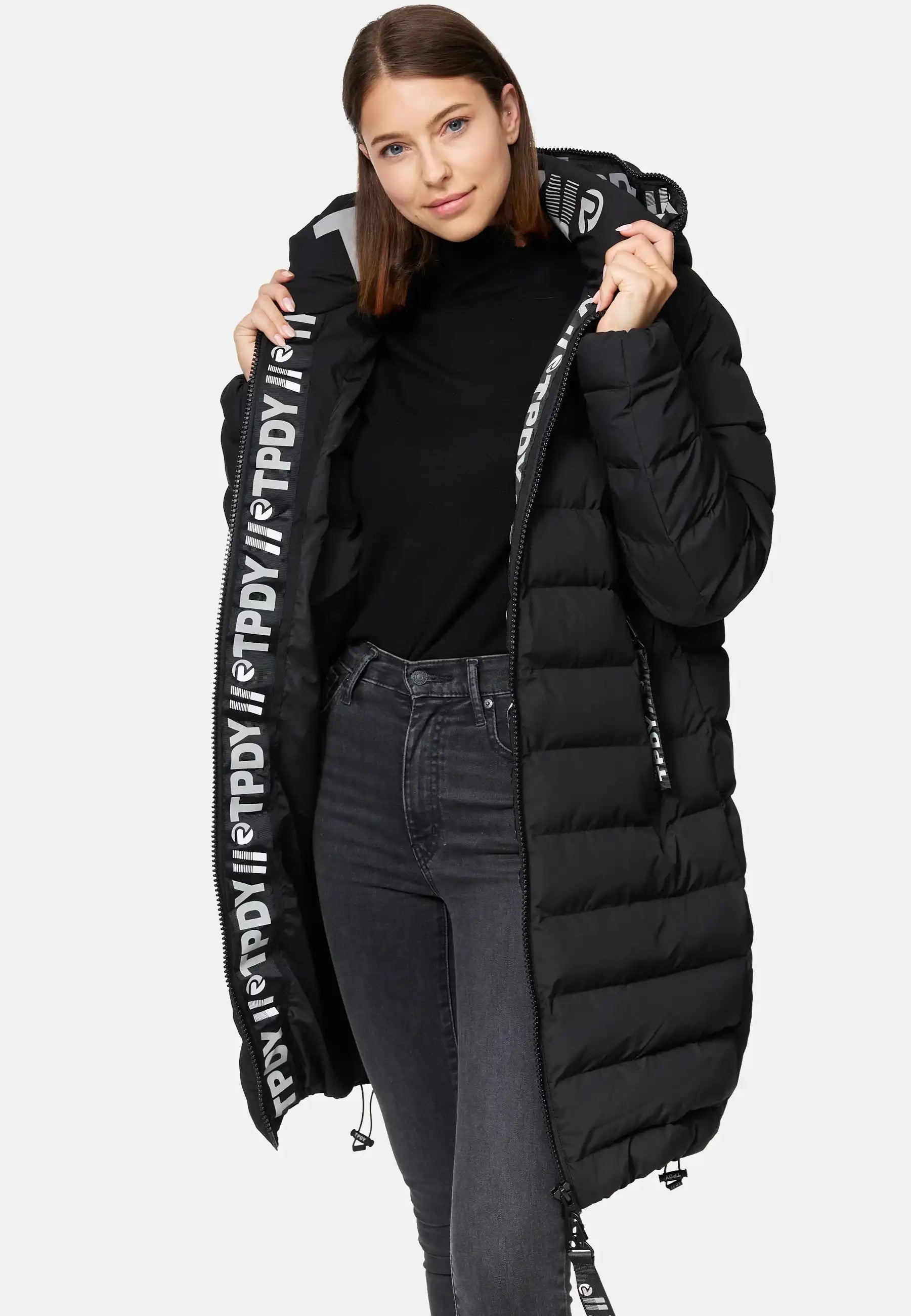 Eine Frau modelliert die trueprodigy Chester F Lange Pufferjacke mit Logo-ein langer, schwarzer, wasserabweisender Damenparka mit auffälligem weißen Logo am Reißverschluss. Sie trägt dazu einen schwarzen Rollkragenpullover und dunkle Jeans.