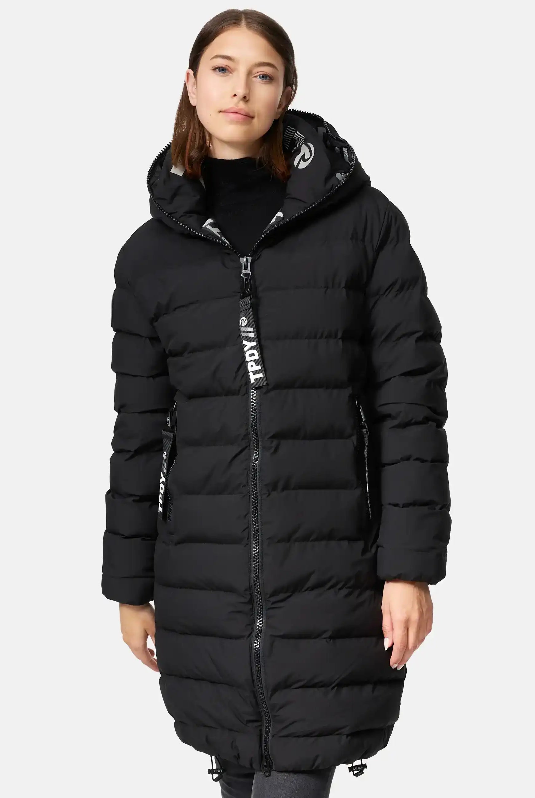 Eine Frau trägt die trueprodigy Chester F Lange Pufferjacke mit Logo-eine wasserabweisende lange schwarze Steppjacke mit Kapuze-sowie einen schwarzen Rollkragenpullover und dunkle Hose, stehend vor weißem Hintergrund.