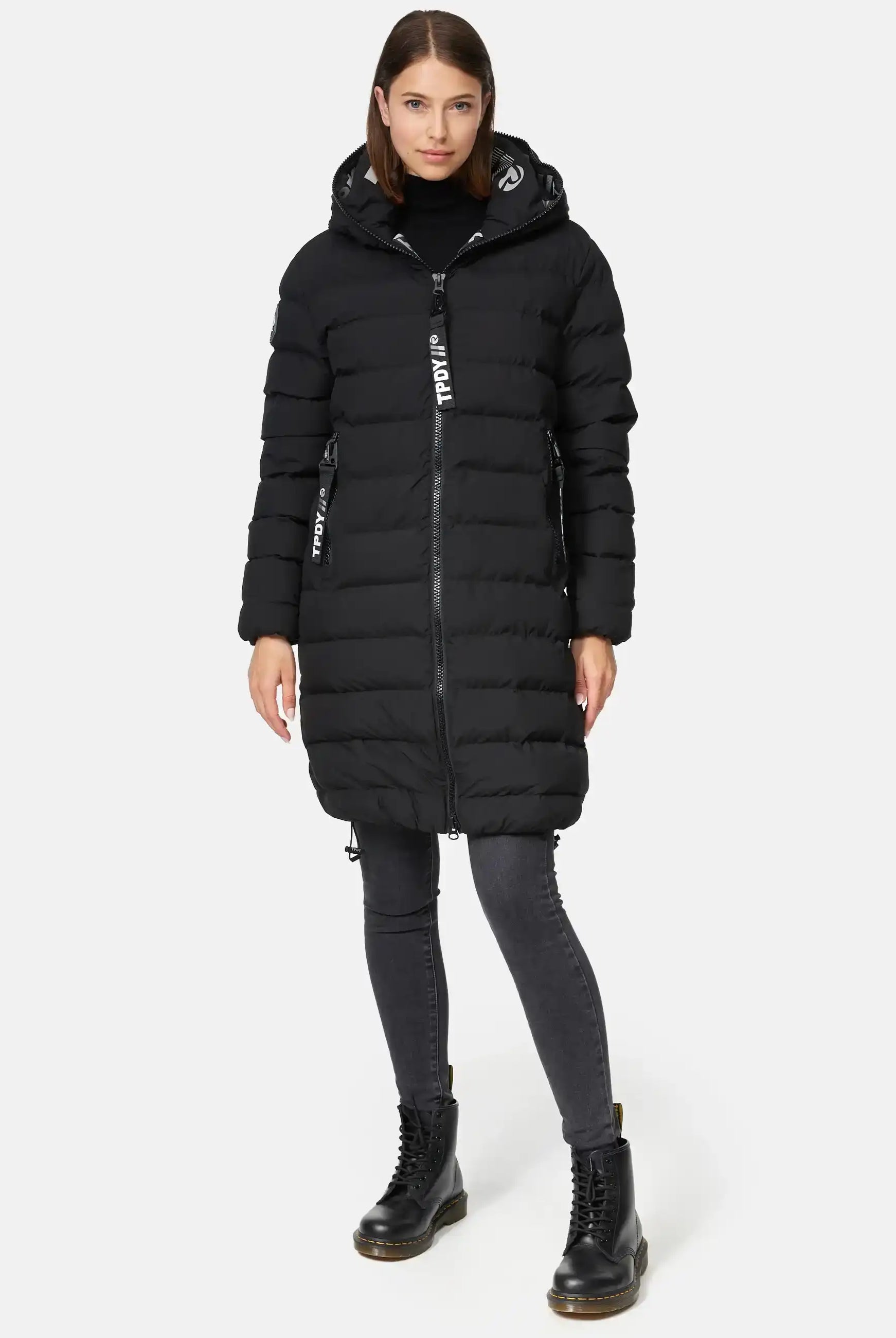 Eine Person trägt die Chester F Lange Pufferjacke mit Logo von trueprodigy, schwarze Jeans und schwarze Schnürstiefel. Die gesteppte Jacke ist geschlossen, die Person blickt neutral nach vorne vor weißem Hintergrund.