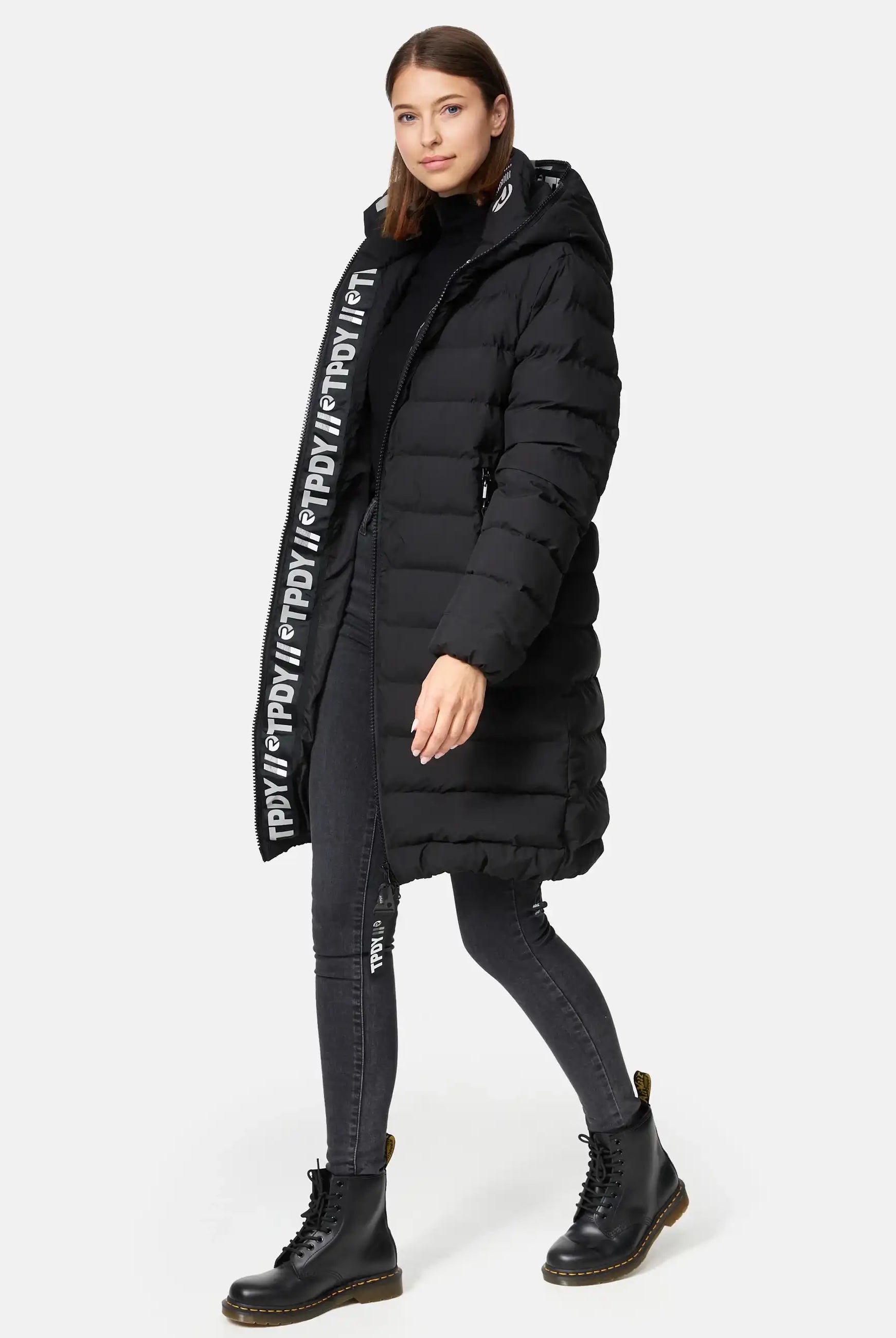 Eine Frau in der trueprodigy Chester F Lange Pufferjacke mit Logo, schwarzen Skinny Jeans und schwarzen Schnürstiefeln geht vor einem schlichten hellen Hintergrund nach vorne.