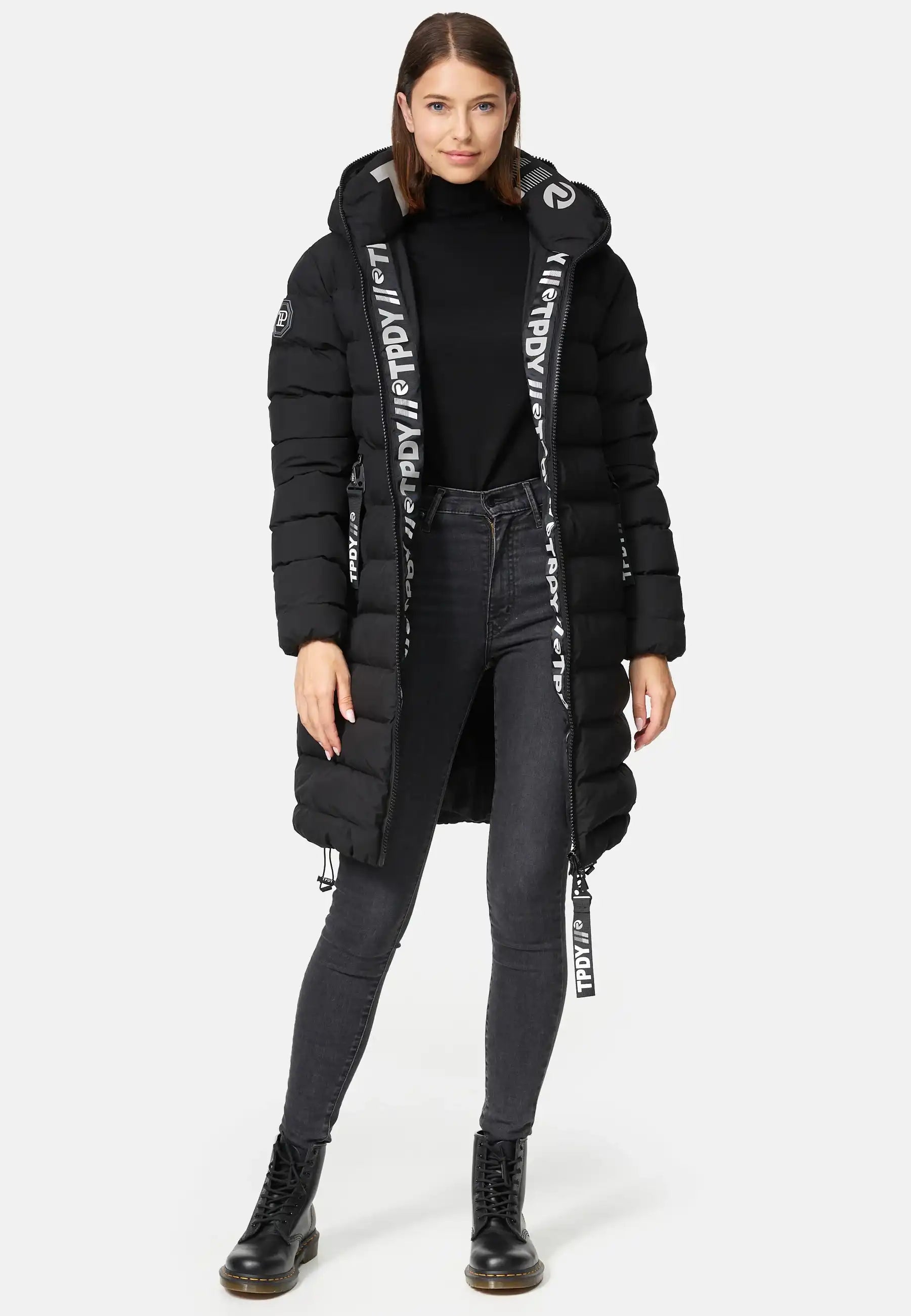 Eine Frau trägt die Chester F Lange Pufferjacke mit Logo von trueprodigy im schwarzen Design mit gesteppten Details und großer Kapuzenbeschriftung, kombiniert mit schwarzem Rollkragenpullover, dunkler Skinny-Jeans und Schnürboots.