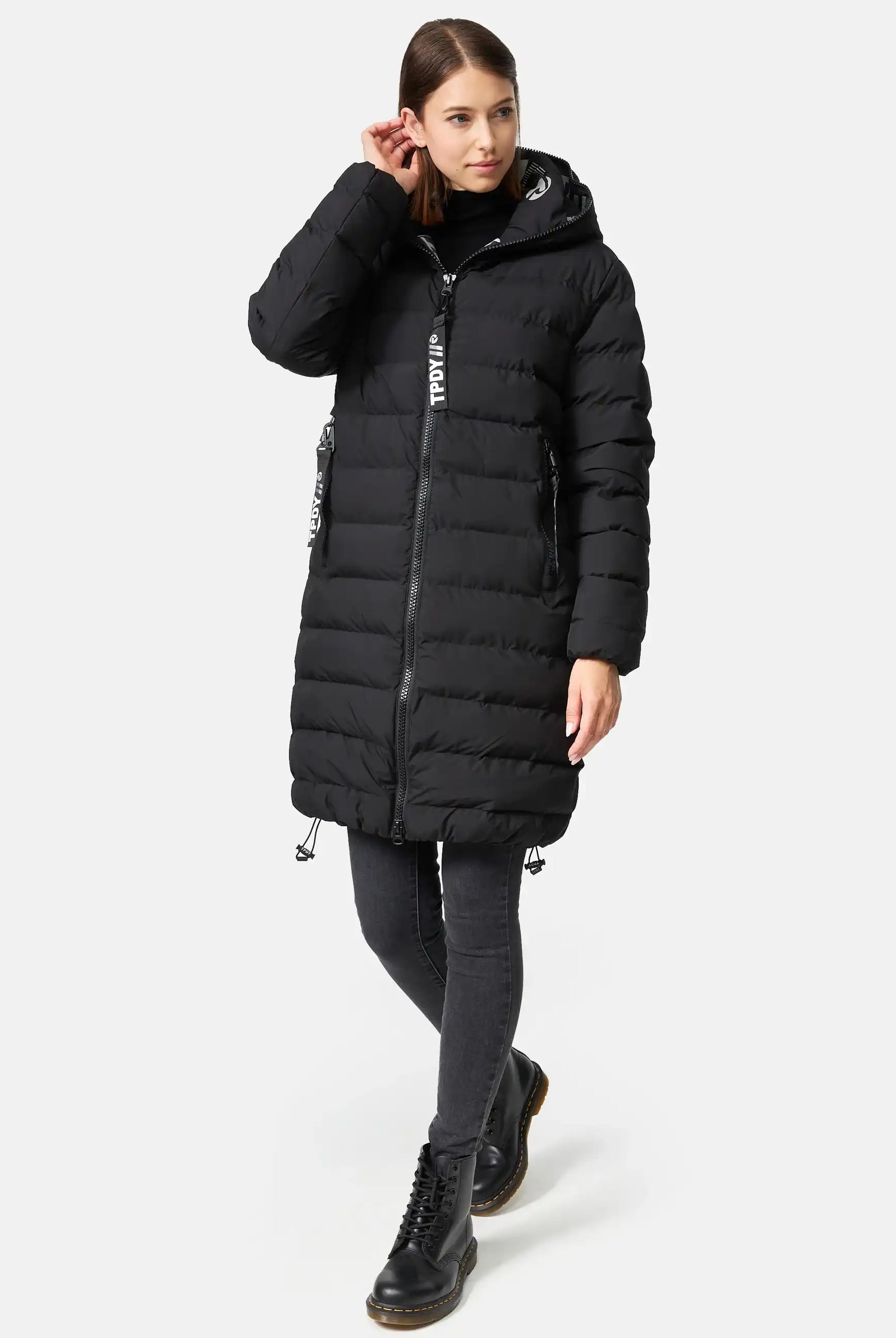 Eine Frau trägt die Chester F Lange Pufferjacke mit Logo von trueprodigy, schwarze Skinny-Jeans und wasserabweisende, schwarze Schnürstiefel. Sie steht vor weißem Hintergrund, berührt ihr Haar und blickt leicht zur Seite.