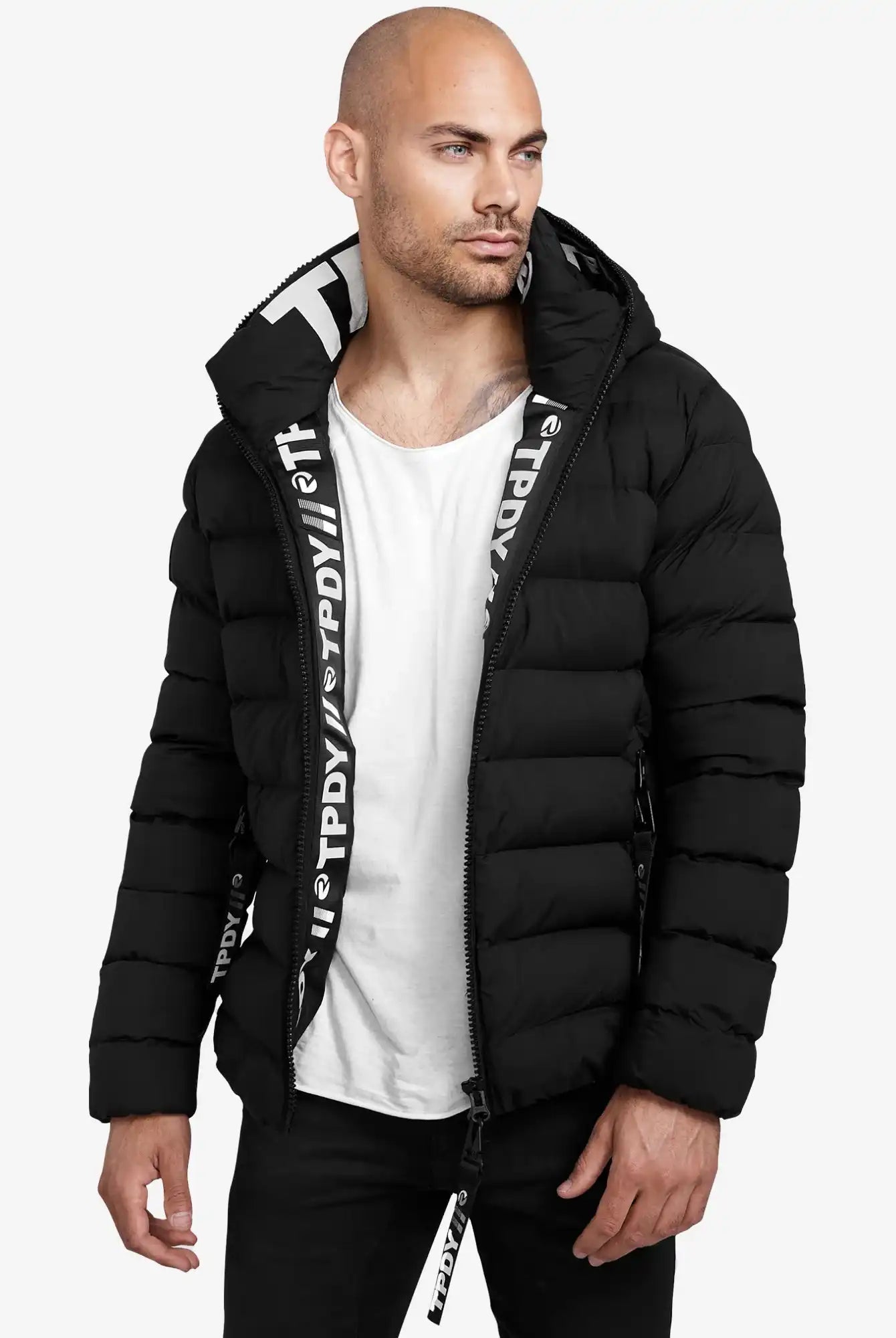Ein Mann trägt eine schwarze Neo Kurze Pufferjacke mit Logo von trueprodigy mit Kapuze, dazu ein weißes T-Shirt und schwarze Hose, steht vor weißem Hintergrund und blickt nach links.