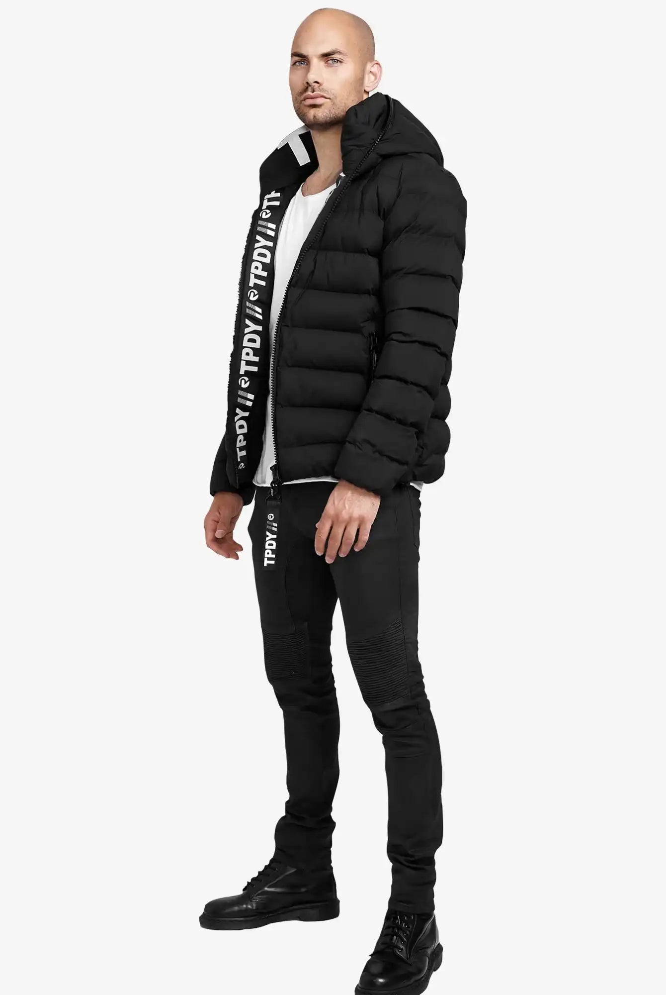 Ein Mann steht vor einem schlichten Hintergrund und trägt die trueprodigy Neo Kurze Pufferjacke mit Logo, eine schwarze Hose mit weißem Text, ein weißes Hemd und schwarze Stiefel. Er steht leicht seitlich und hat die Arme entspannt.
