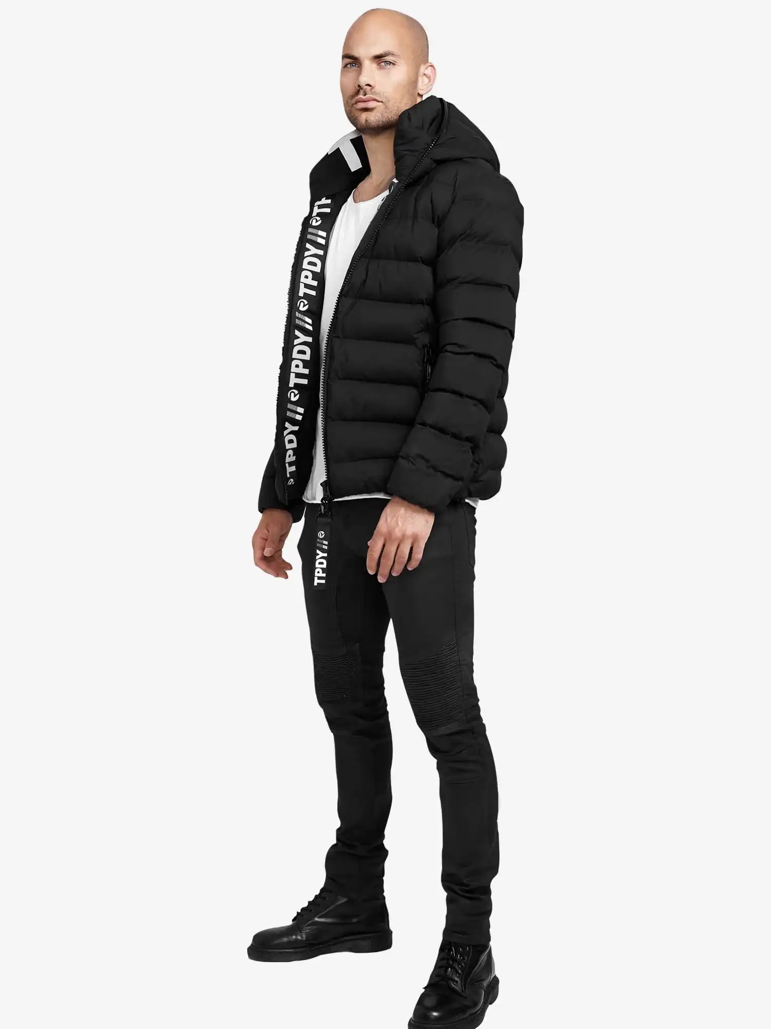 Ein Mann steht vor einem schlichten Hintergrund und trägt die trueprodigy Neo Kurze Pufferjacke mit Logo, eine schwarze Hose mit weißem Text, ein weißes Hemd und schwarze Stiefel. Er steht leicht seitlich und hat die Arme entspannt.