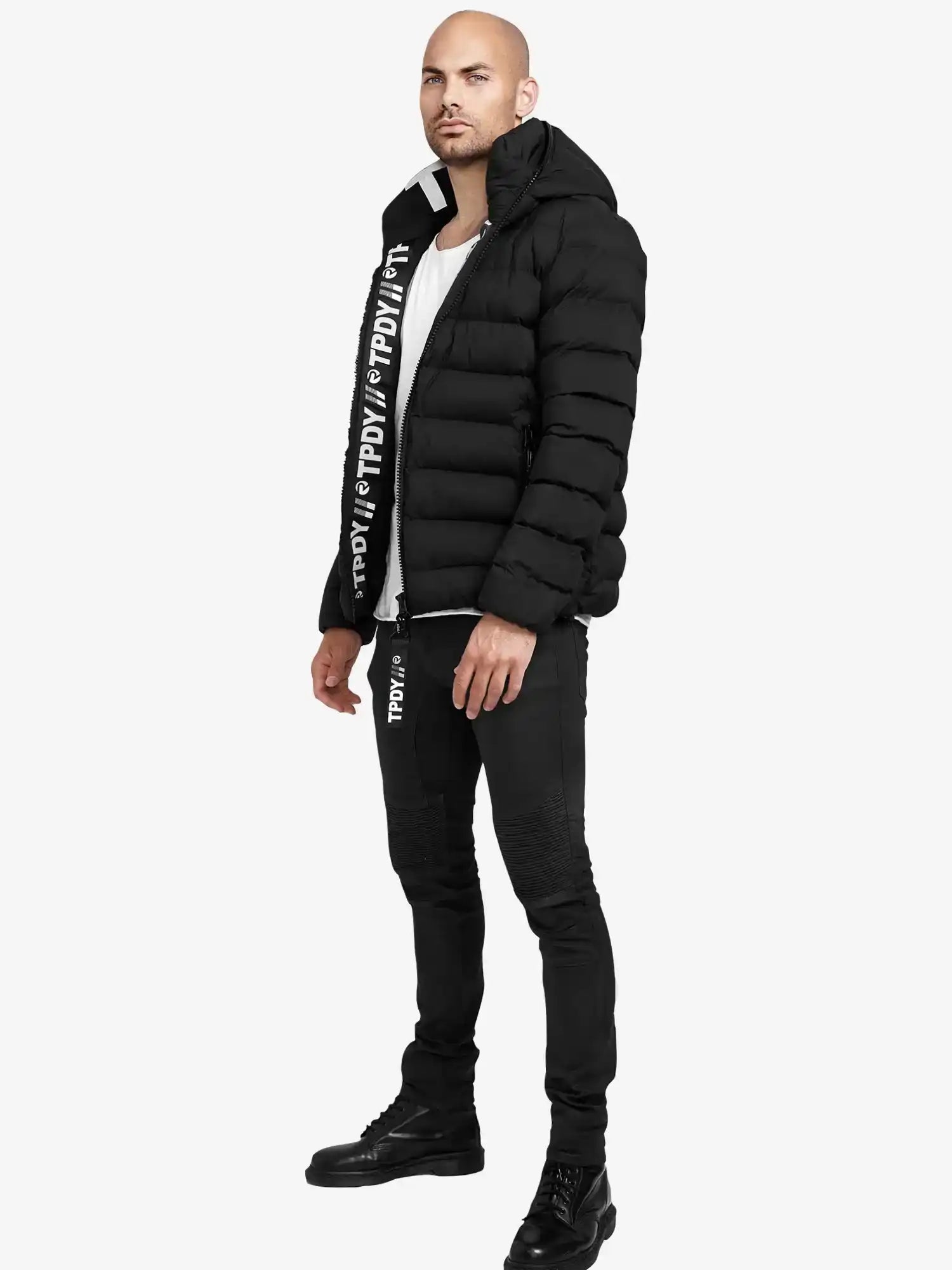 Ein Mann steht vor einem schlichten Hintergrund und trägt die trueprodigy Neo Kurze Pufferjacke mit Logo, eine schwarze Hose mit weißem Text, ein weißes Hemd und schwarze Stiefel. Er steht leicht seitlich und hat die Arme entspannt.