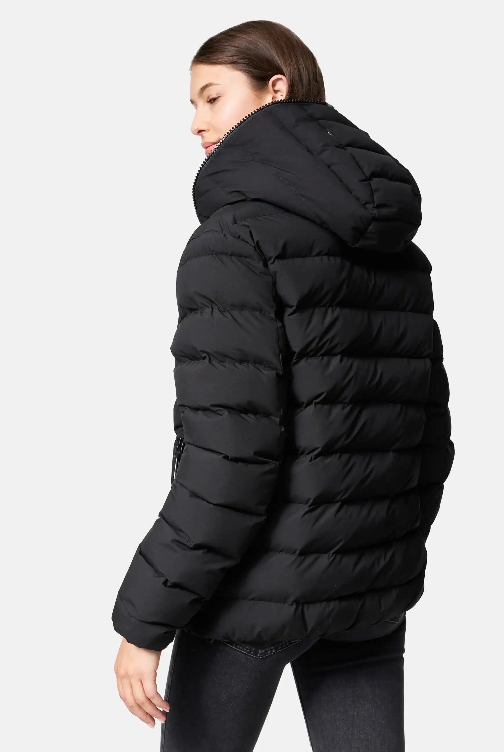 Eine Person mit kurzen braunen Haaren, die die trueprodigy Neo F Kurze Pufferjacke mit Logo trägt, steht vor einem schlichten hellen Hintergrund und schaut leicht über ihre Schulter.