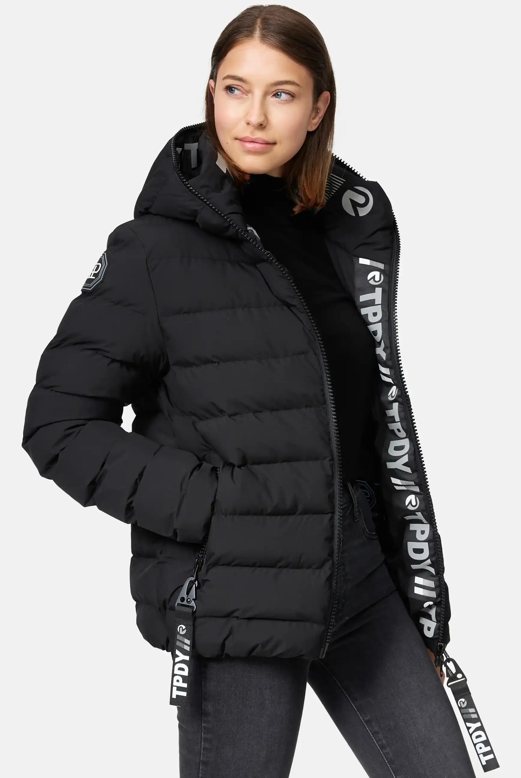 Eine Frau mit glattem braunem Haar trägt die trueprodigy Neo F Kurze Pufferjacke mit Logo, eine wasserabweisende Damenjacke mit fester Kapuze und fettem weißem Logotext auf dem Reißverschluss und den Kordelzügen, über einem schwarzen Rollkragenpullover. Sie posiert mit einer Hand in ihrer Tasche.