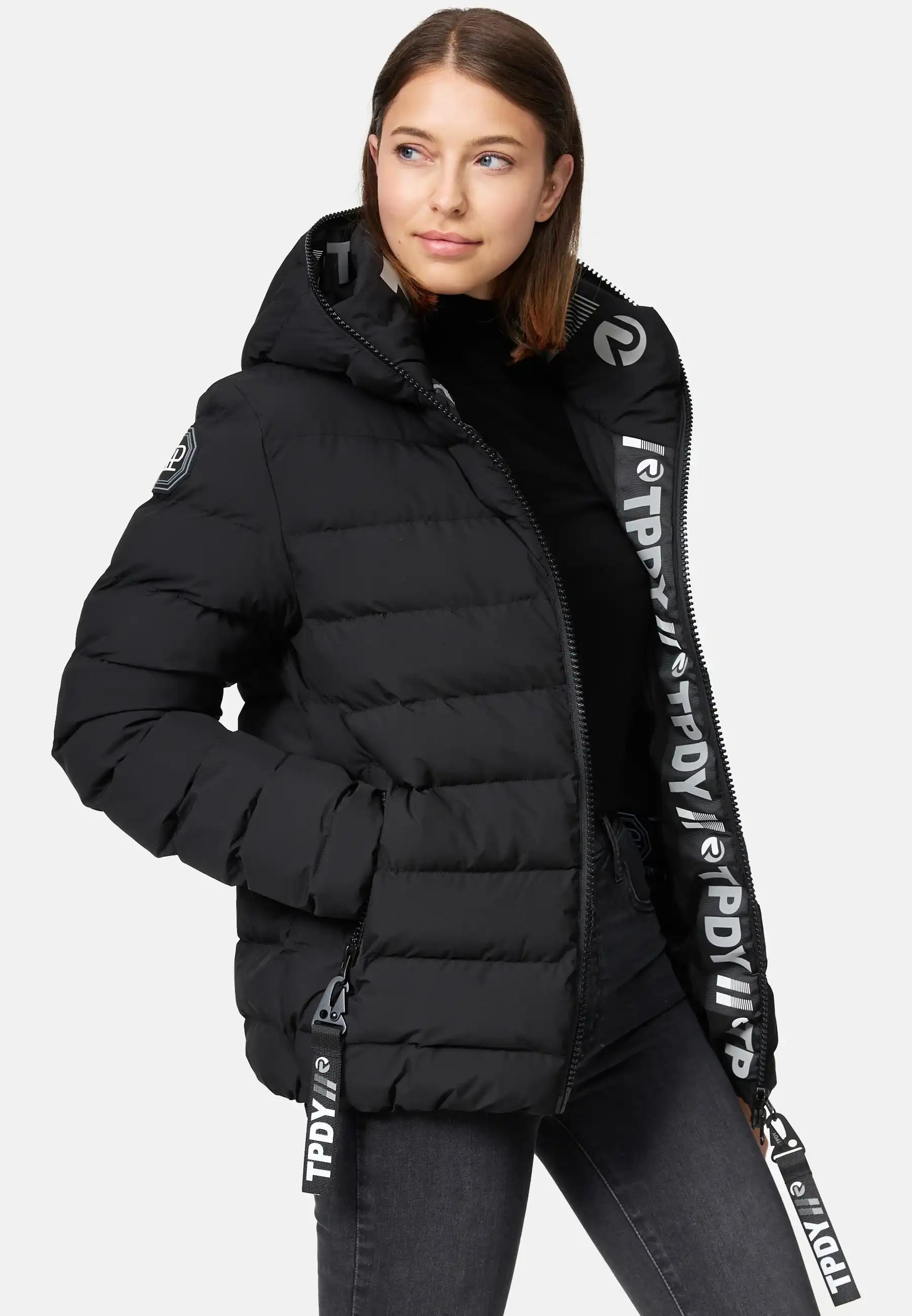 Eine Frau mit glattem braunem Haar trägt die trueprodigy Neo F Kurze Pufferjacke mit Logo, eine wasserabweisende Damenjacke mit fester Kapuze und fettem weißem Logotext auf dem Reißverschluss und den Kordelzügen, über einem schwarzen Rollkragenpullover. Sie posiert mit einer Hand in ihrer Tasche.