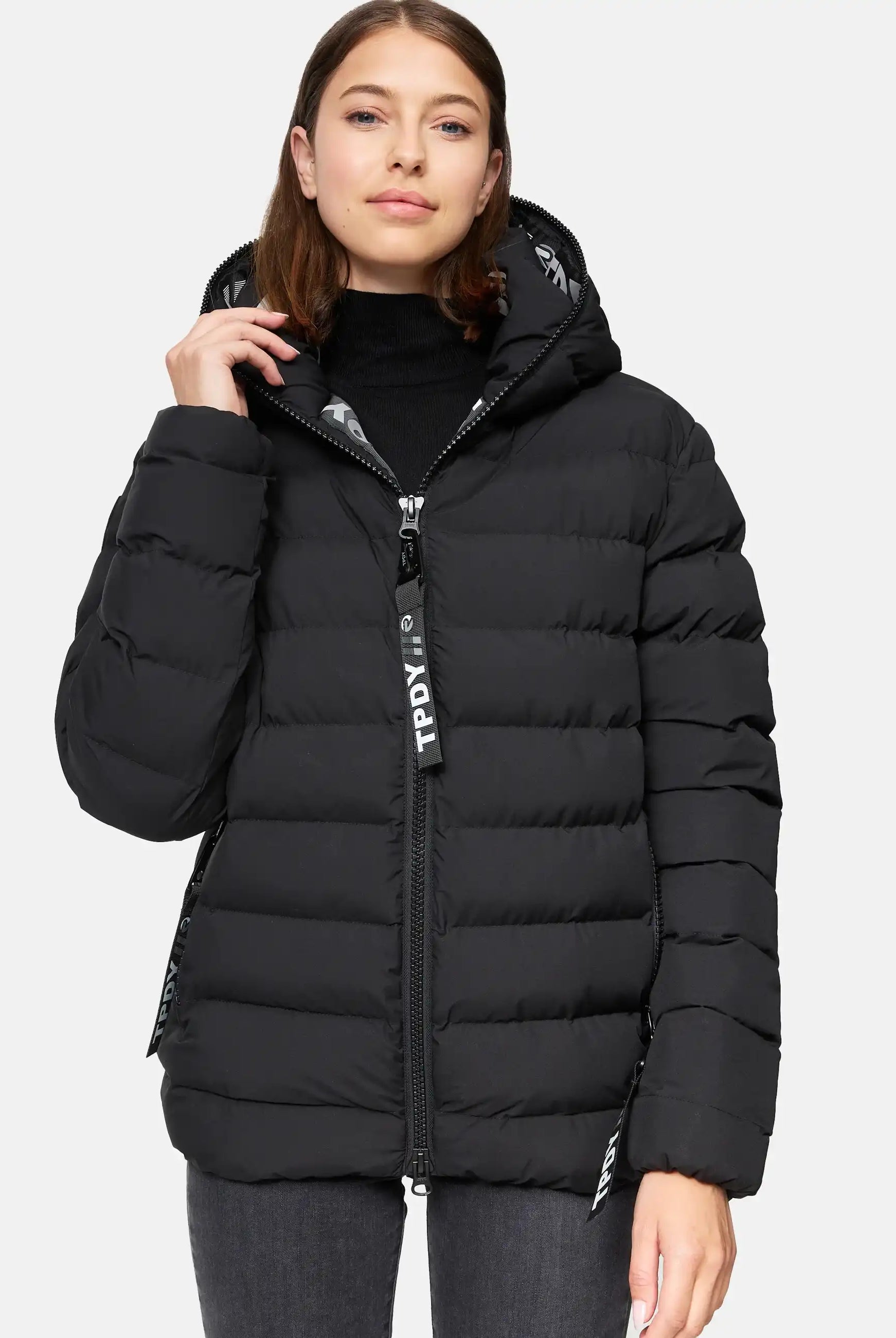 Eine Frau trägt die trueprodigy Neo F Kurze Pufferjacke mit Logo, eine schwarze, wasserabweisende Damenjacke mit Kapuze, über einem schwarzen Rollkragenpullover und grauen Jeans. Sie schaut nach vorne und hält den Reißverschluss. Die Jacke hat große Pull-Tags mit weißem Logo-Text.