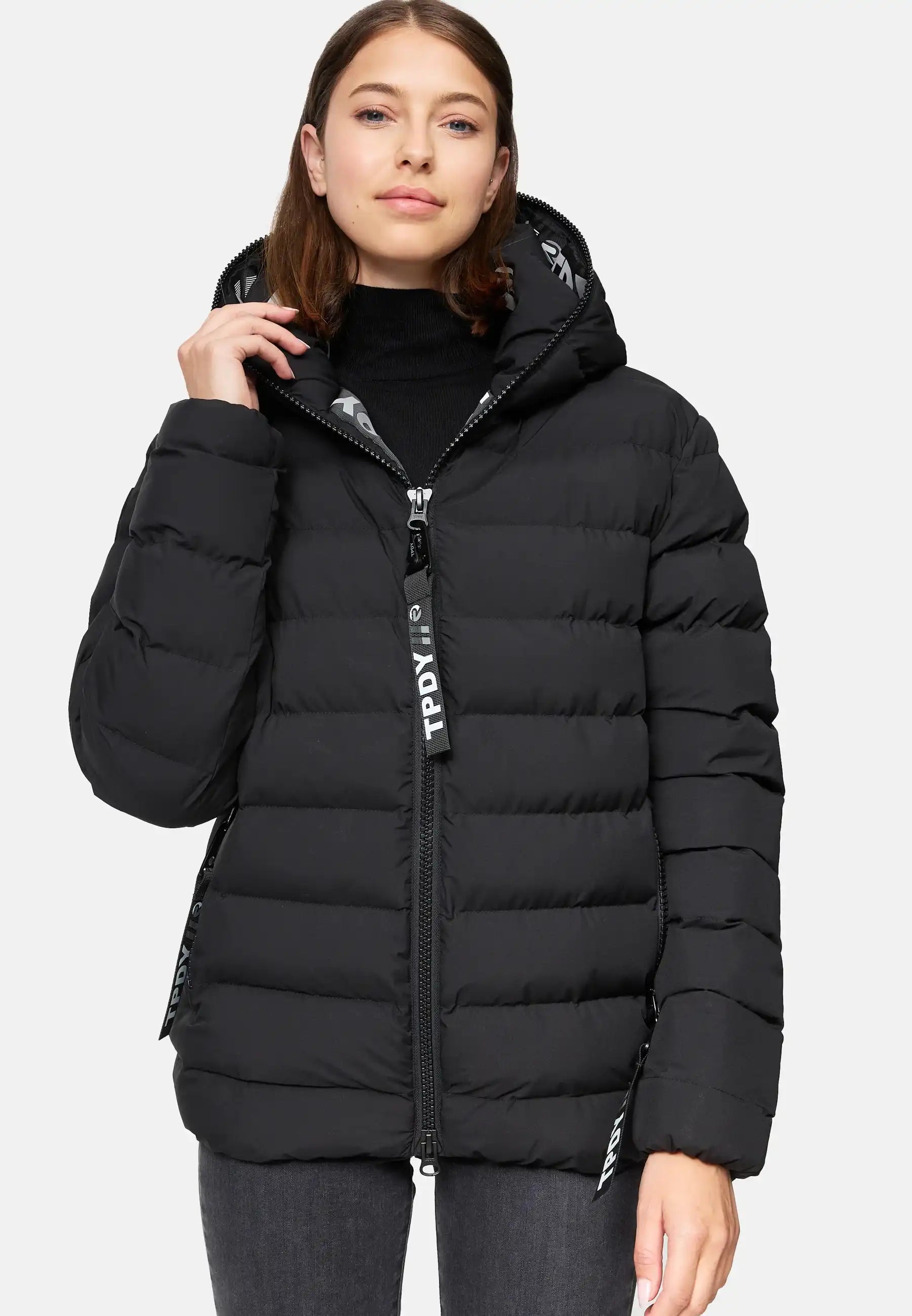 Eine Frau trägt die trueprodigy Neo F Kurze Pufferjacke mit Logo, eine schwarze, wasserabweisende Damenjacke mit Kapuze, über einem schwarzen Rollkragenpullover und grauen Jeans. Sie schaut nach vorne und hält den Reißverschluss. Die Jacke hat große Pull-Tags mit weißem Logo-Text.