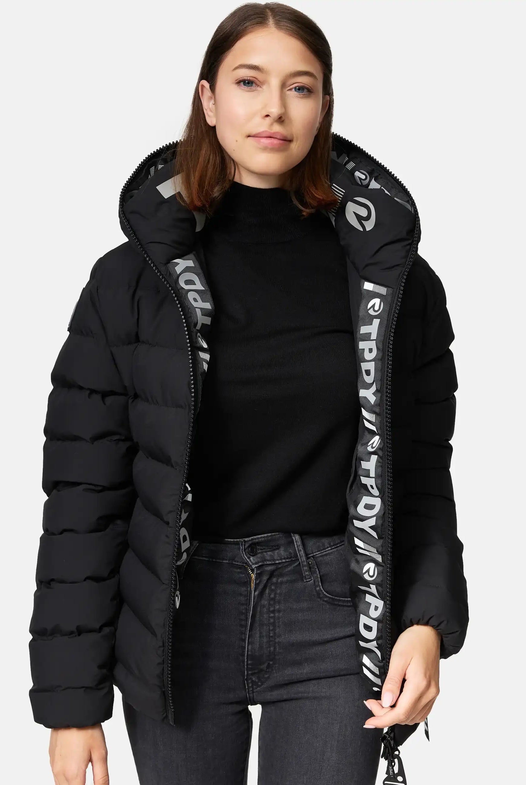 Eine Frau modelliert die trueprodigy Neo F kurze Pufferjacke mit Logo in Schwarz, mit fester Kapuze und wasserabweisendem Material. Sie trägt darunter einen schwarzen Rollkragen, dunkle Jeans und zeigt das gemusterte Innenfutter der offenen Jacke.