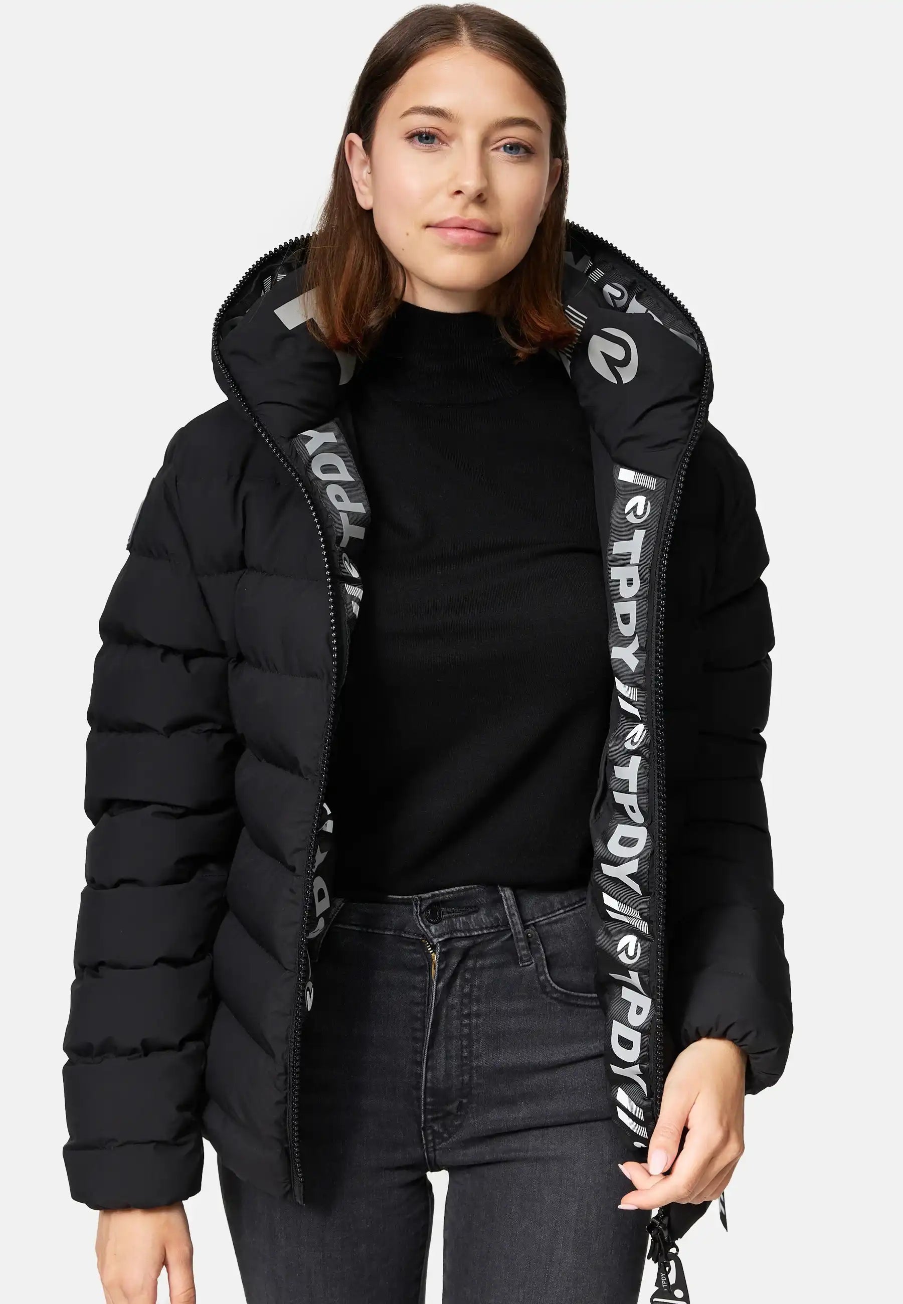 Eine Frau modelliert die trueprodigy Neo F kurze Pufferjacke mit Logo in Schwarz, mit fester Kapuze und wasserabweisendem Material. Sie trägt darunter einen schwarzen Rollkragen, dunkle Jeans und zeigt das gemusterte Innenfutter der offenen Jacke.