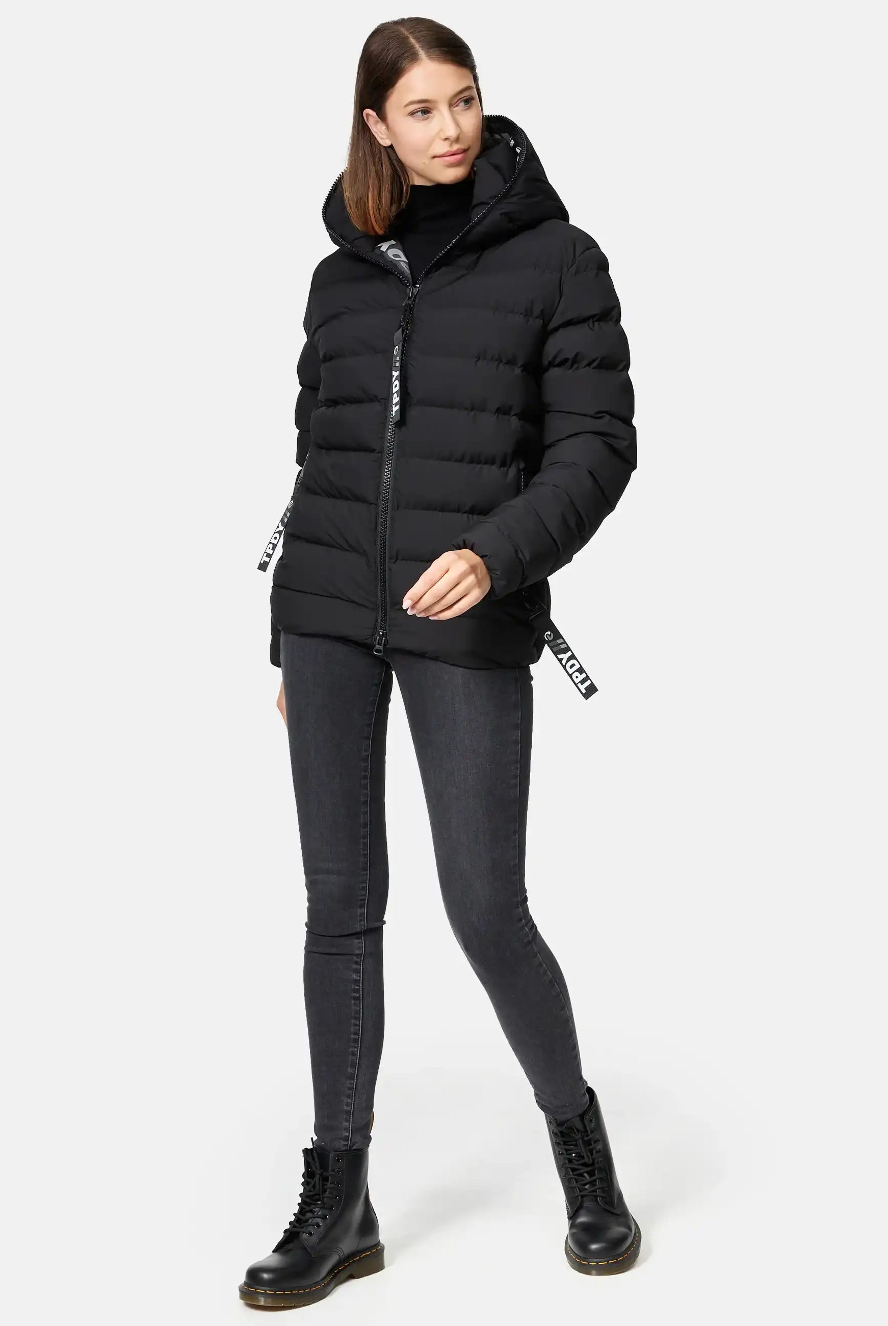 Eine Frau mit glattem braunem Haar steht leicht rechts, trägt die trueprodigy Neo F Kurze Pufferjacke mit Logo, einen schwarzen Rollkragenpullover, dunkelgraue Skinny Jeans und schwarze Schnürstiefel, eine Hand in der Jackentasche.