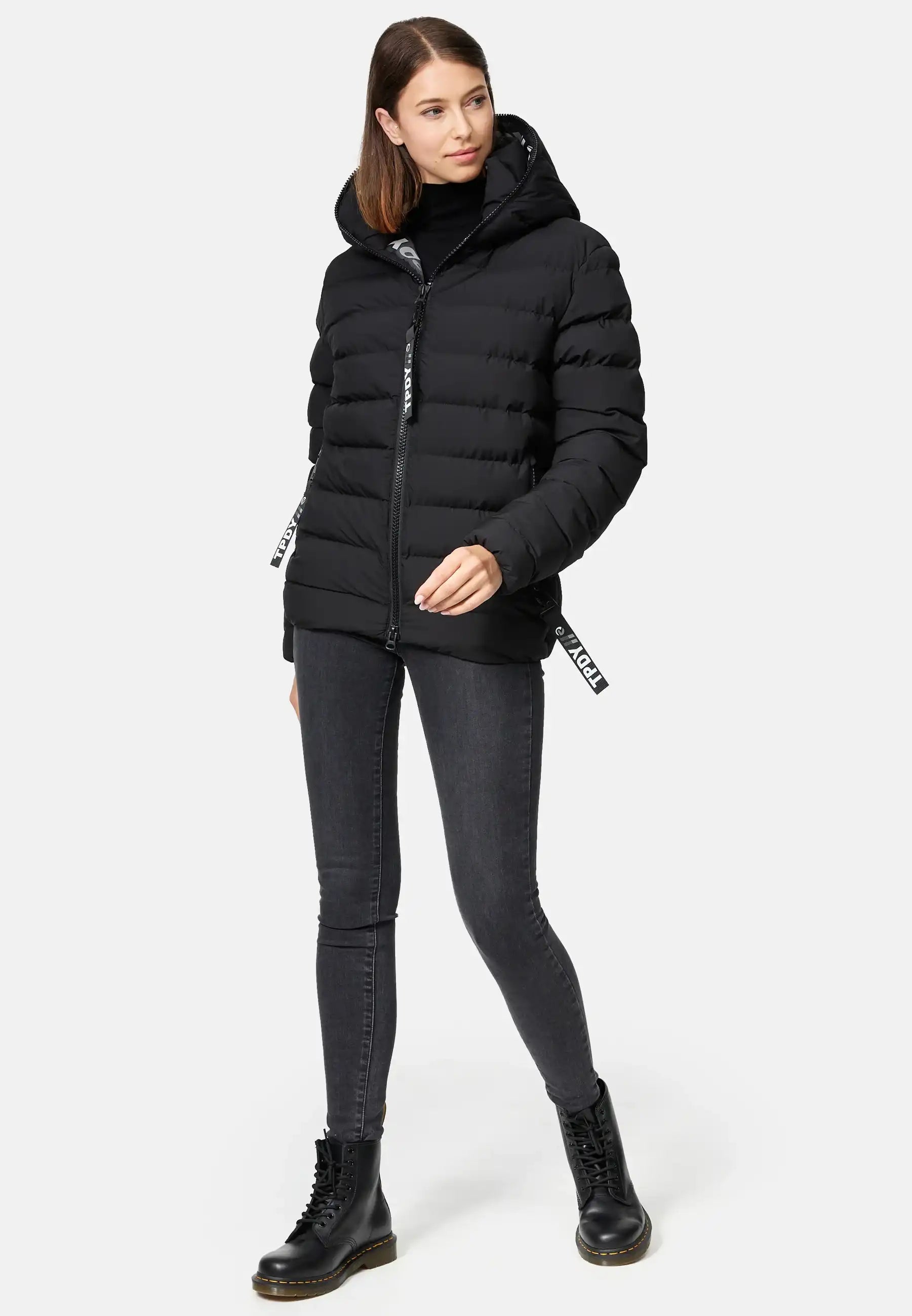 Eine Frau mit glattem braunem Haar steht leicht rechts, trägt die trueprodigy Neo F Kurze Pufferjacke mit Logo, einen schwarzen Rollkragenpullover, dunkelgraue Skinny Jeans und schwarze Schnürstiefel, eine Hand in der Jackentasche.