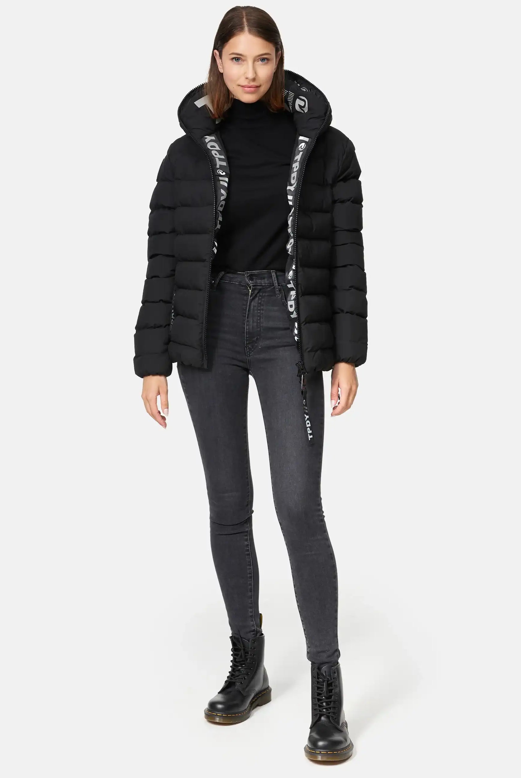 Eine Frau trägt die Neo F Kurze Pufferjacke mit Logo von trueprodigy, kombiniert mit schwarzem Rollkragenpullover, grauen Skinny-Jeans und schwarzen Schnürstiefeln vor hellem Hintergrund.