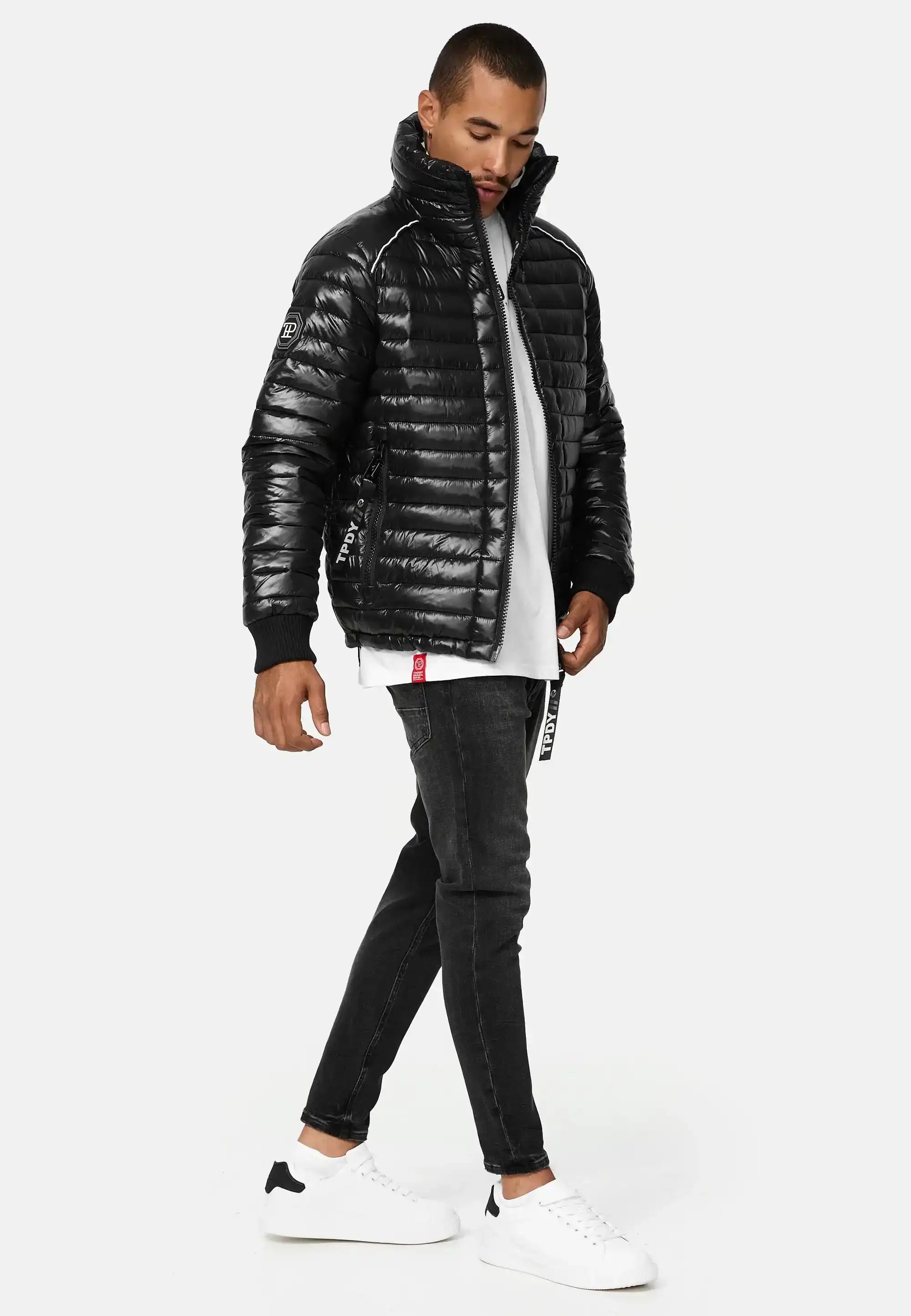 Ein Mann steht vor einem schlichten Hintergrund und trägt die Trueprodigy Corey Steppjacke mit Stehkragen, eine glänzende schwarze Pufferjacke, gepaart mit einem weißen T-Shirt, schwarzen Jeans und weißen Turnschuhen. Er schaut mit einer Hand in der Jackentasche nach unten.